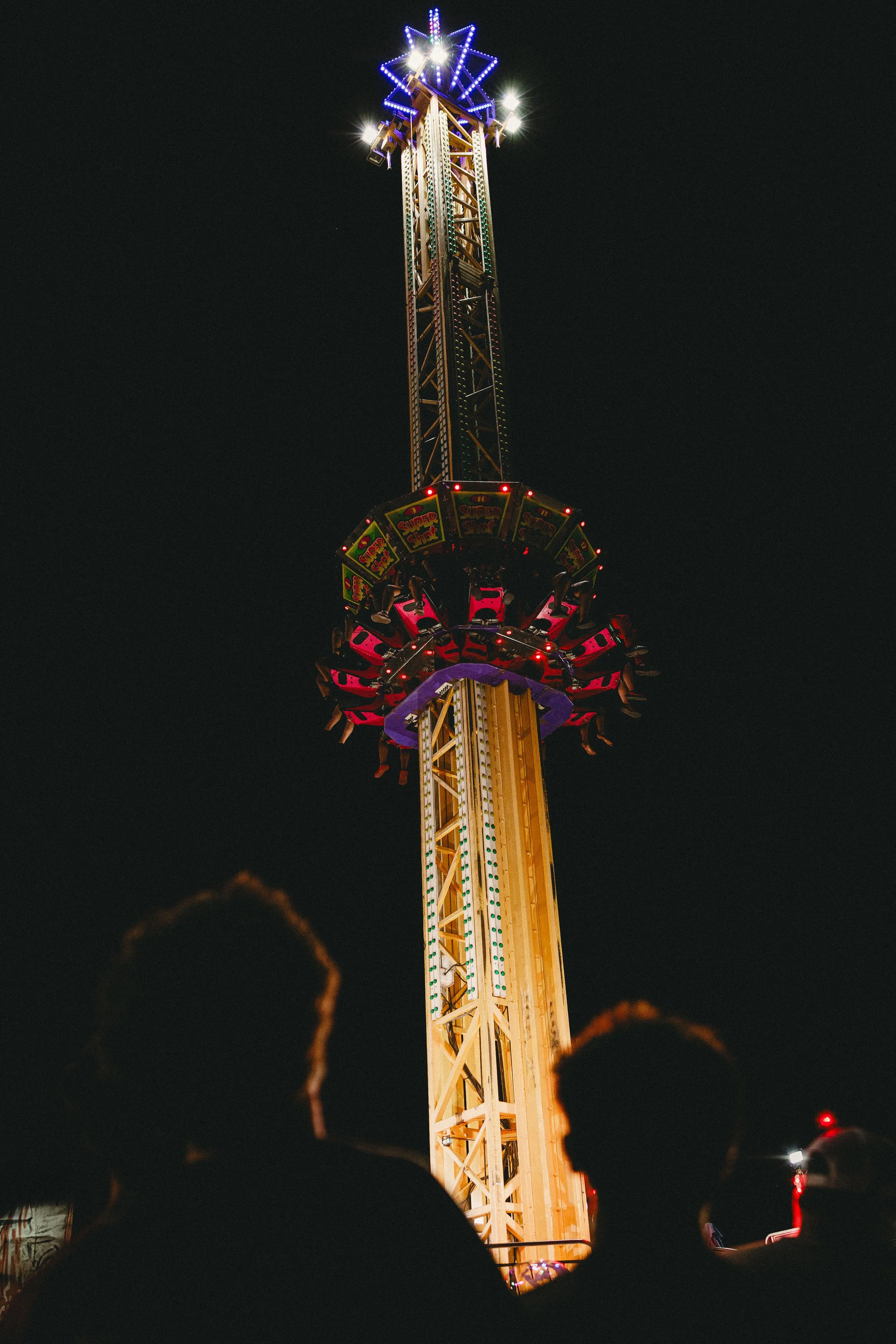 Nighttime view of a tall amusement park drop tower ride illuminated by colorful lights, with three silhouetted people in the foreground. Shot at Fiesta Topeka. 