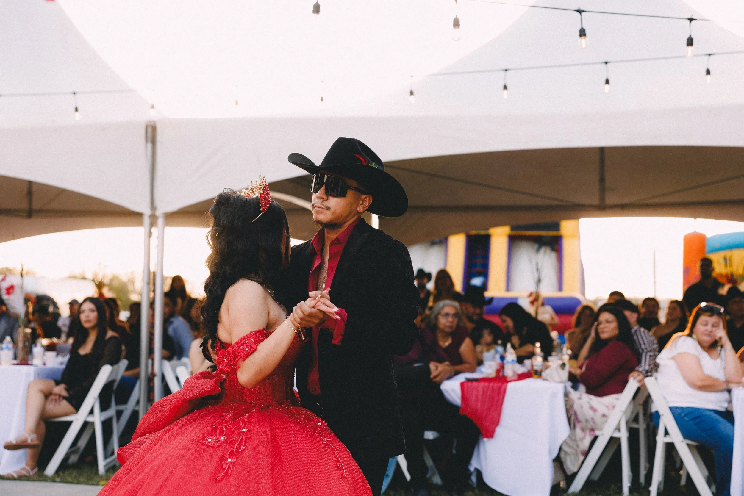 A couple dancing at an outdoor event with white tents and seated guests in the background. The woman is wearing a red ball gown and a tiara, while the man is dressed in black with sunglasses and a hat.