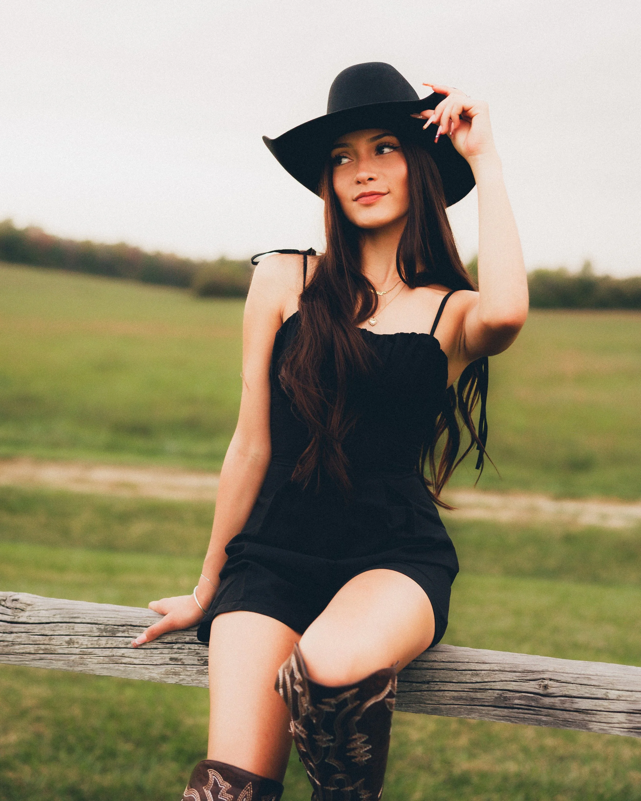 Young woman sitting on a wooden fence outdoors, wearing a black dress, a wide-brimmed black hat, and patterned cowboy boots, in a grassy field.