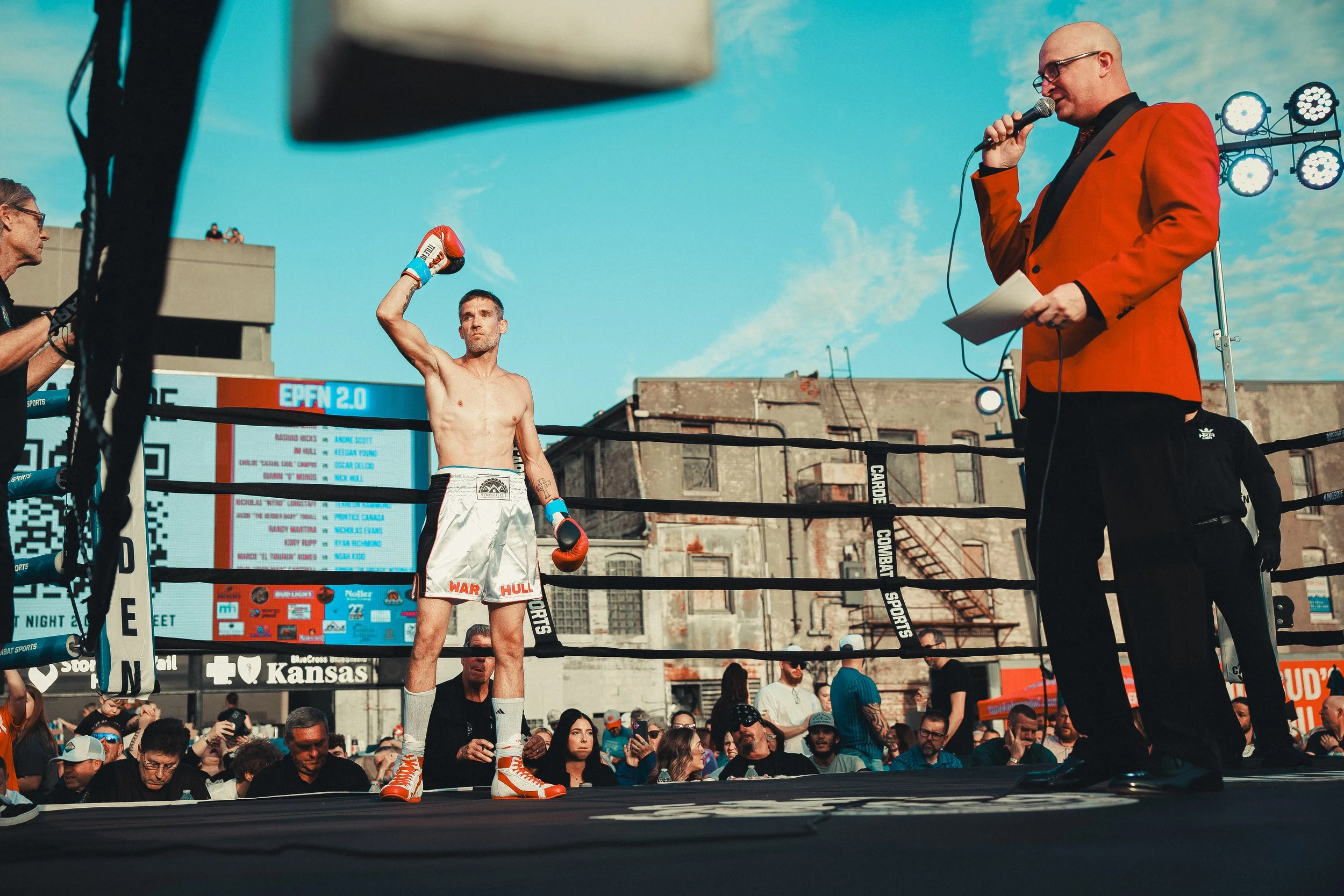 A boxer in white shorts and red boxing gloves stands shirtless inside a ring, raising his right arm in victory while an announcer in an orange blazer speaks into a microphone. The scene takes place outdoors, with a crowd of spectators watching and a 