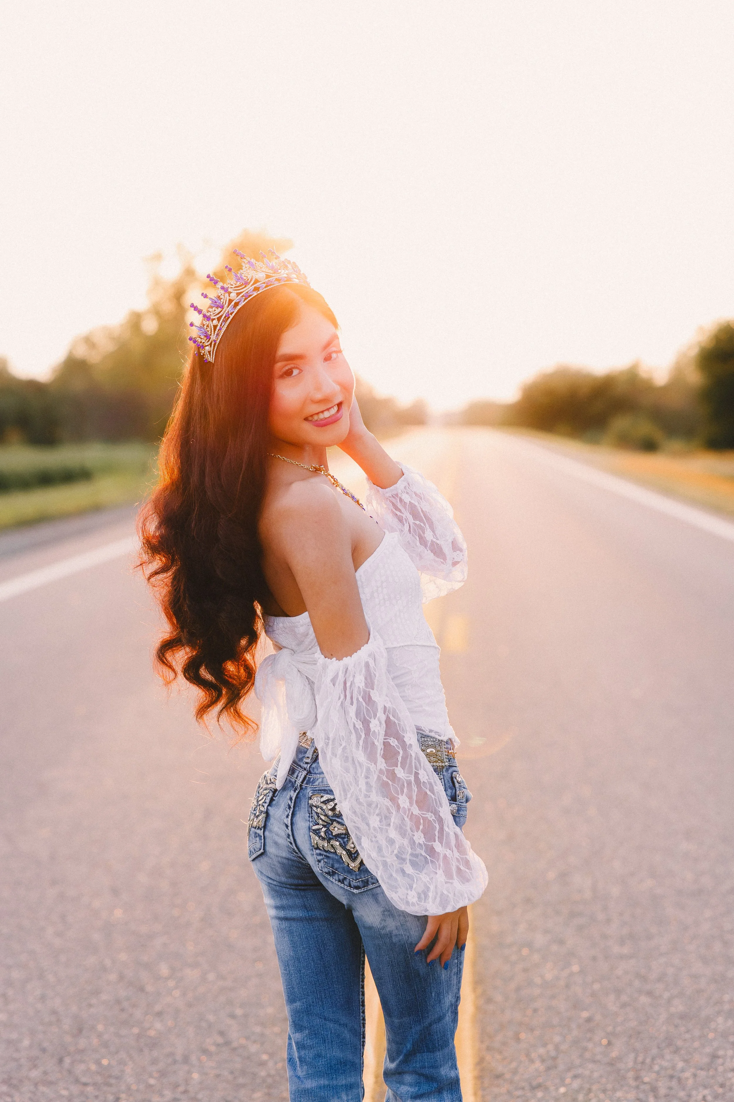Young woman with long, wavy hair wearing a tiara, white lace top, and jeans, standing on a rural road at sunset.
