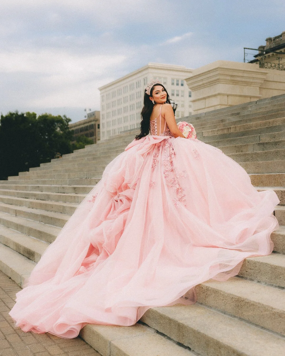A woman in a pink quinceañera dress with a full skirt, lace-up bodice, and tiara, sitting on steps outside a government or historic building, holding a bouquet of pink flowers, smiling at the camera.