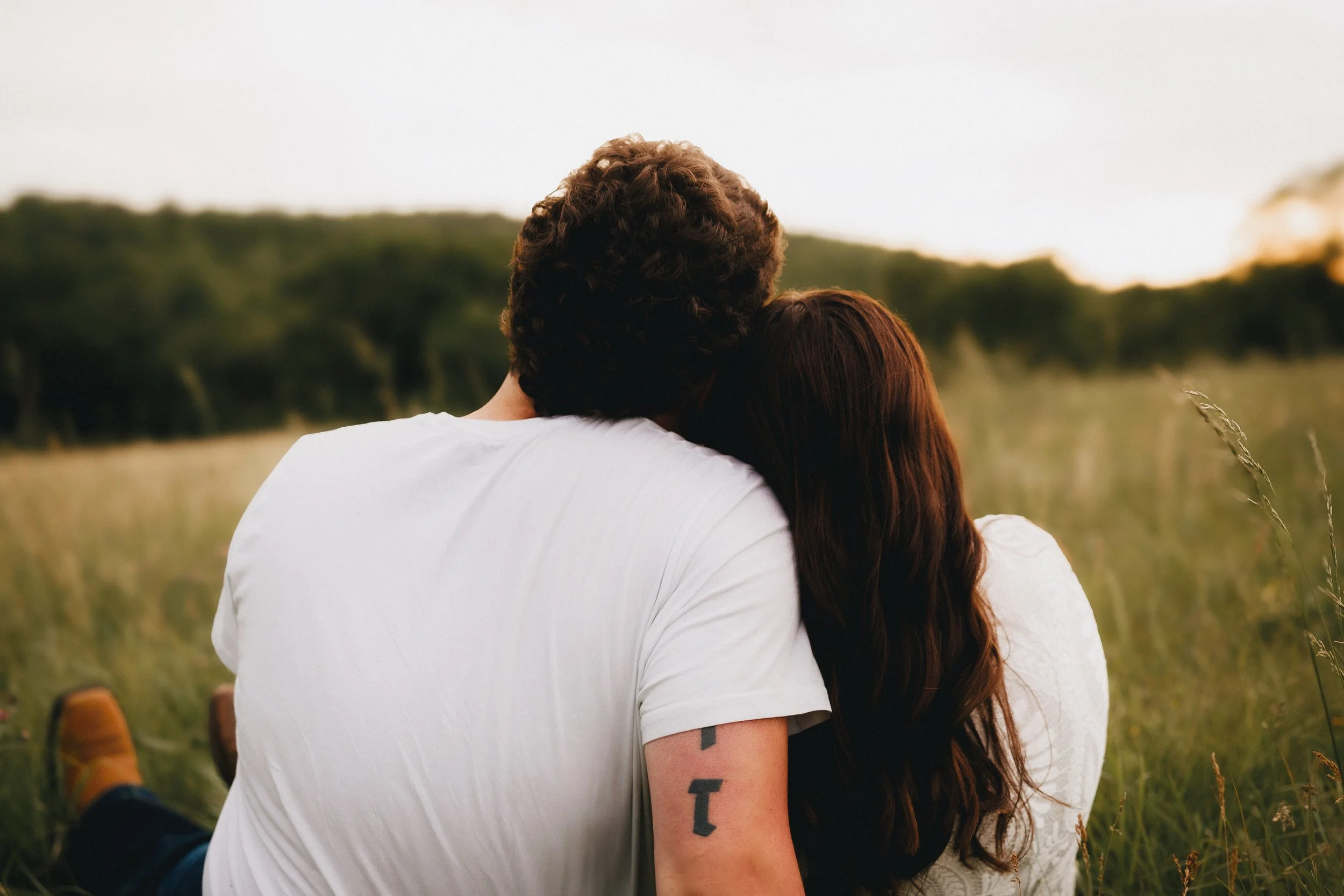 Couple sitting in a grassy field during sunset, leaning into each other for a kiss.