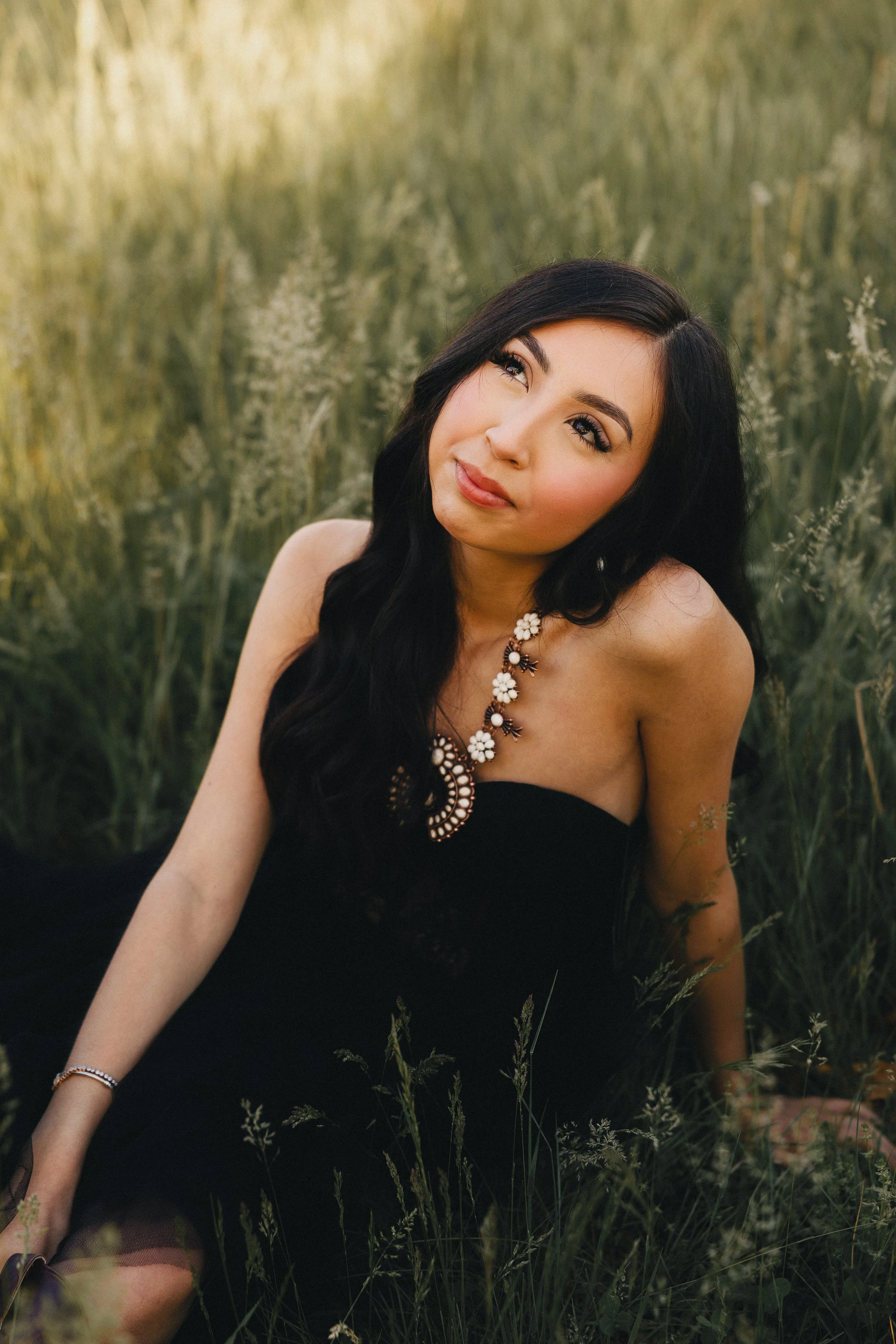 A woman with long black hair sitting in a grassy field, looking thoughtfully upward, wearing a black strapless dress and a statement floral necklace.
