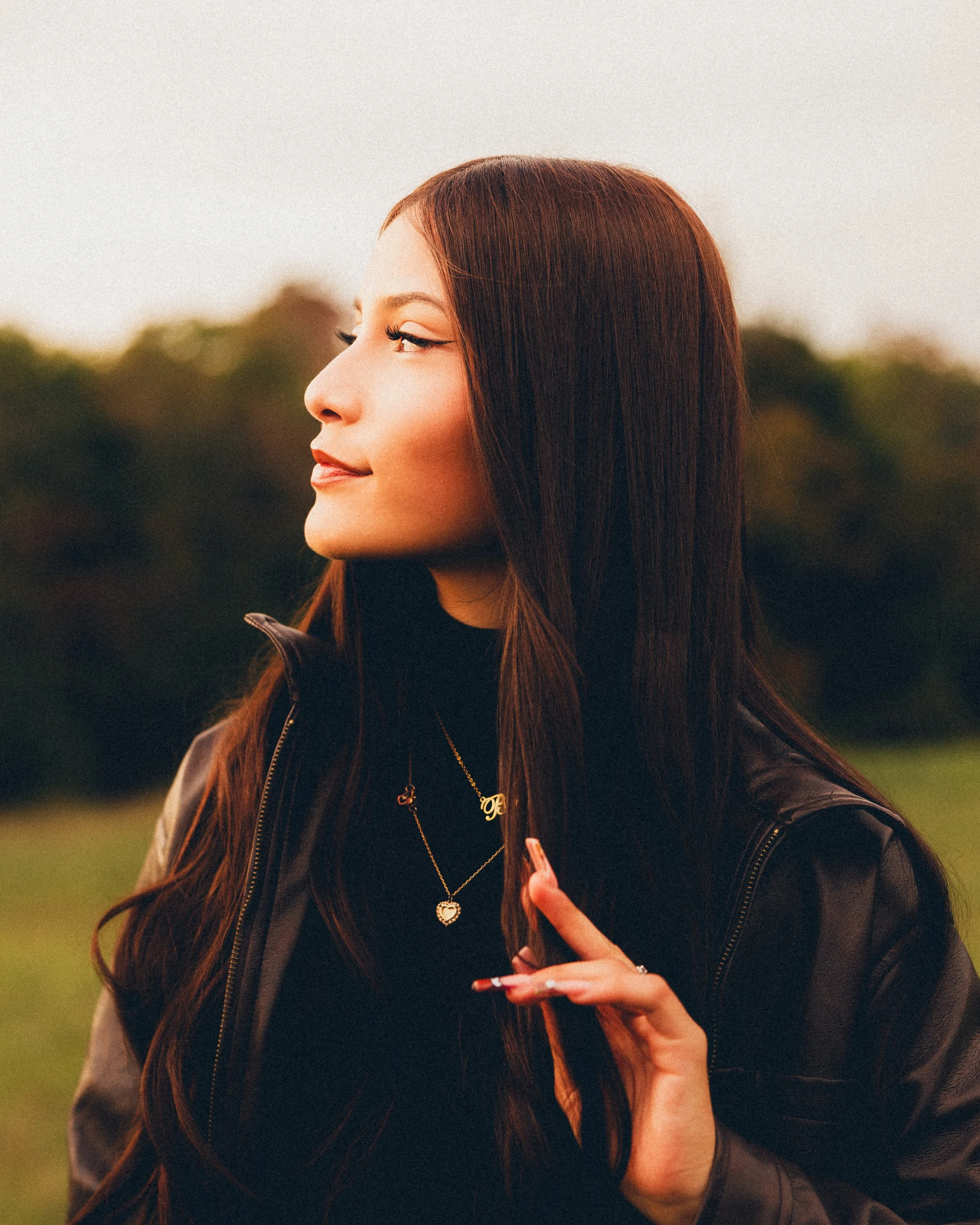 Profile of a young woman with long brown hair, wearing a black turtleneck, layered necklaces, and a black leather jacket, standing outdoors with blurred trees in the background