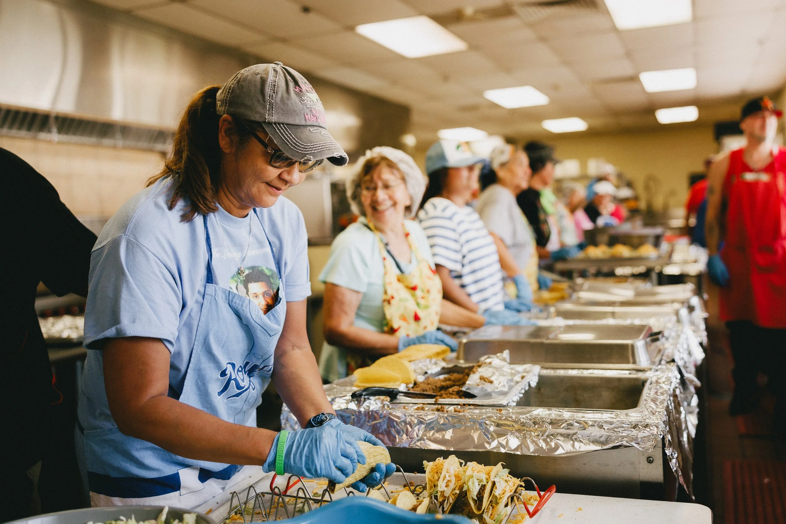 A group of women standing in a food line, wearing aprons and gloves, serving and preparing food at a buffet table in a community kitchen or cafeteria. Shot at Fiesta Topeka.