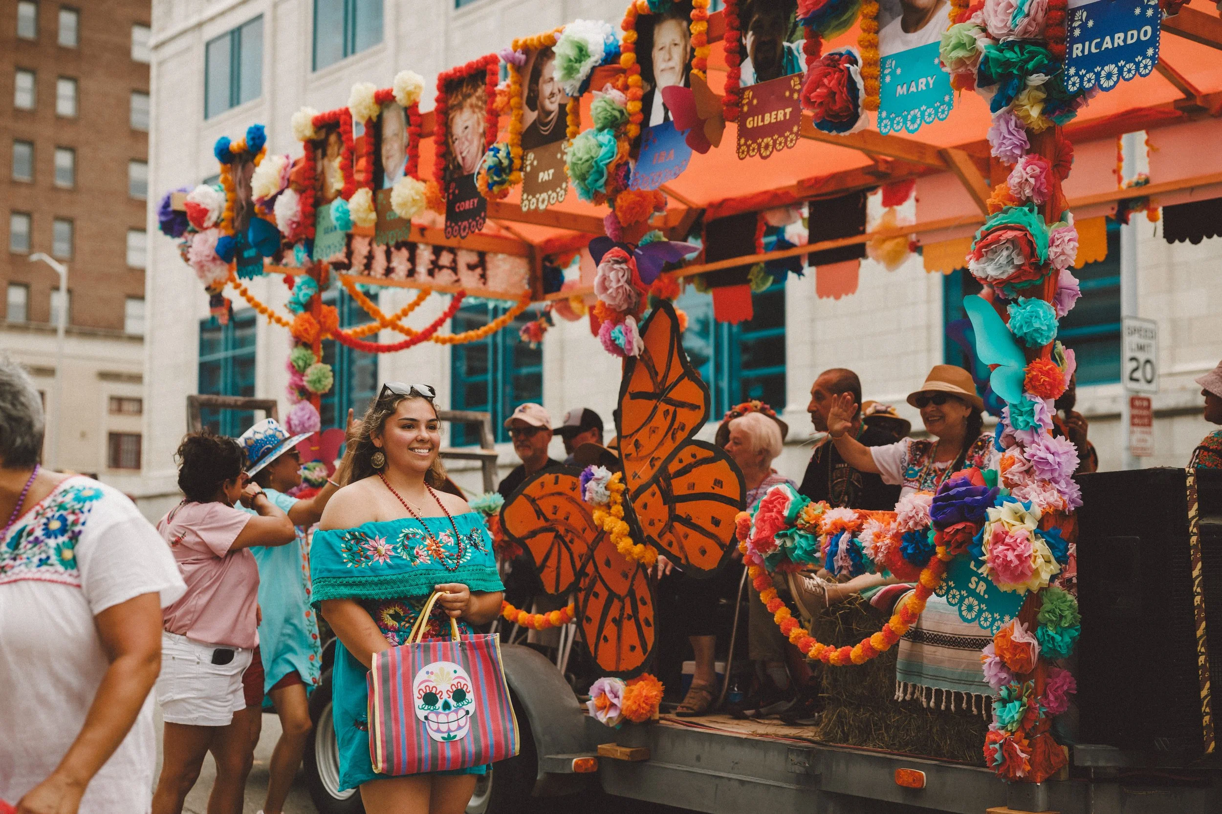 People celebrating on a float decorated with colorful flowers, photos, and butterflies at a parade. Participants are wearing festive attire, including a young woman in a bright blue dress holding a decorated bag. Shot at Fiesta Topeka parade.