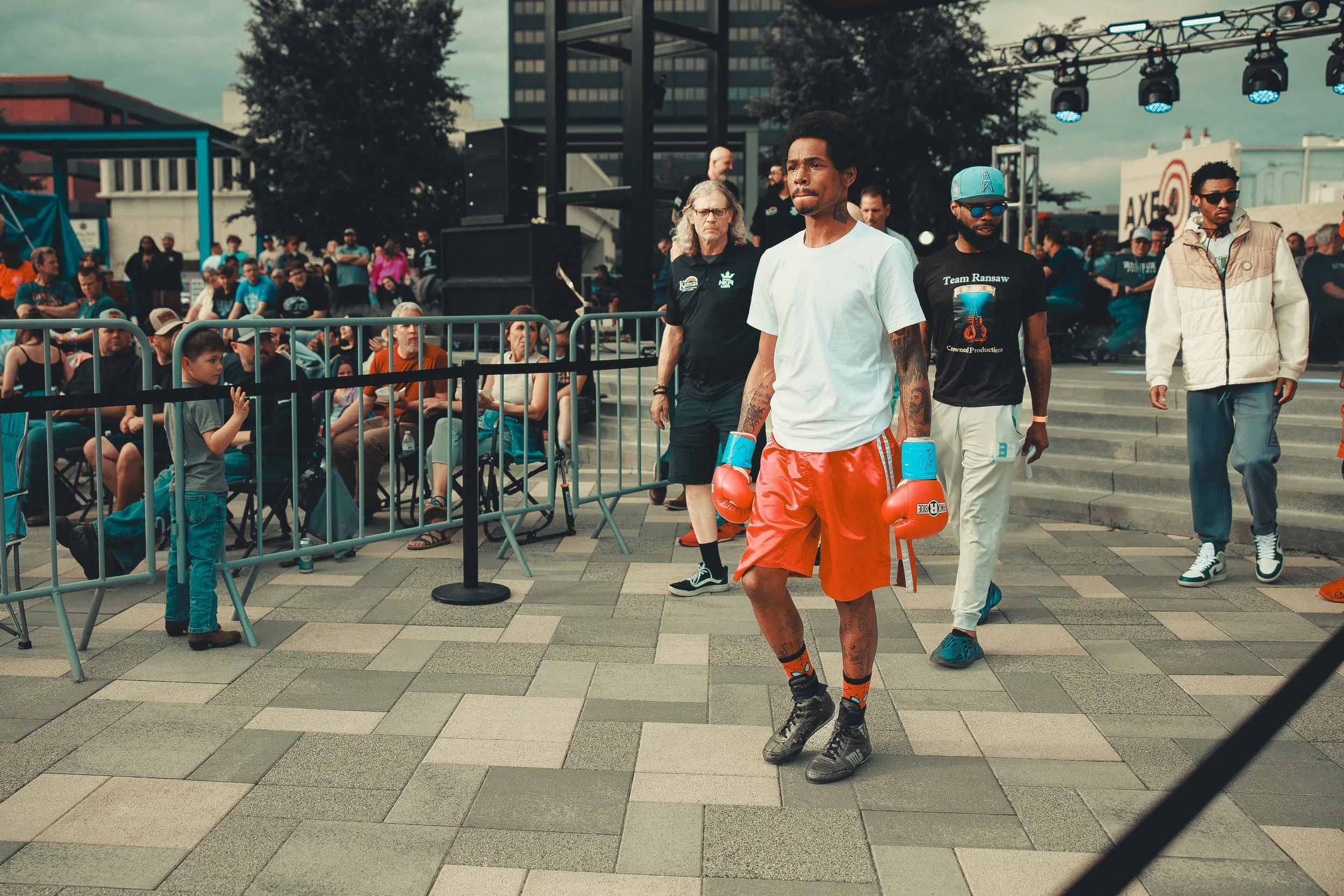 A male boxer in orange shorts and boxing gloves stands in the ring surrounded by a crowd of spectators and onlookers at an outdoor event. Shot at Evergy Plaza in Topeka, Kansas. 
