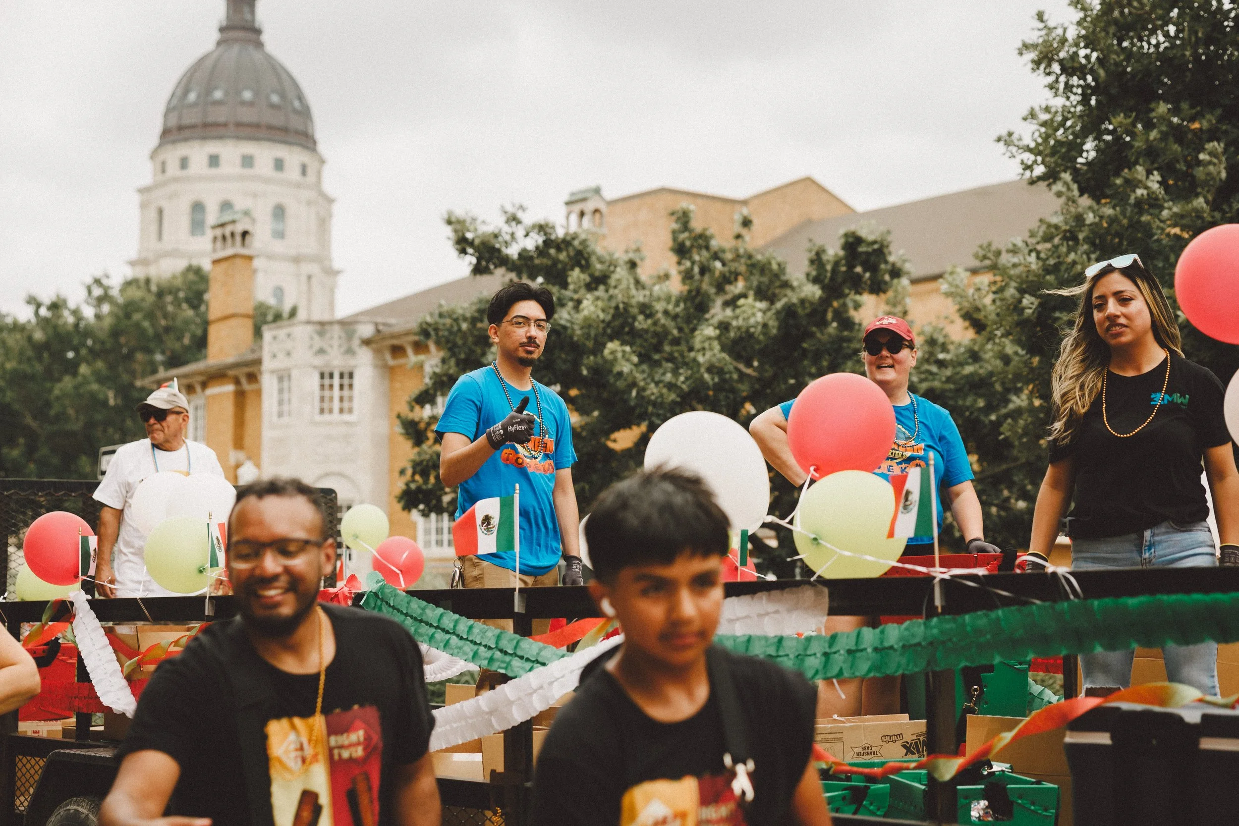 People participating in a float parade with Mexican flags and balloons, with a historic building and trees in the background. Shot at Fiesta Topeka parade.