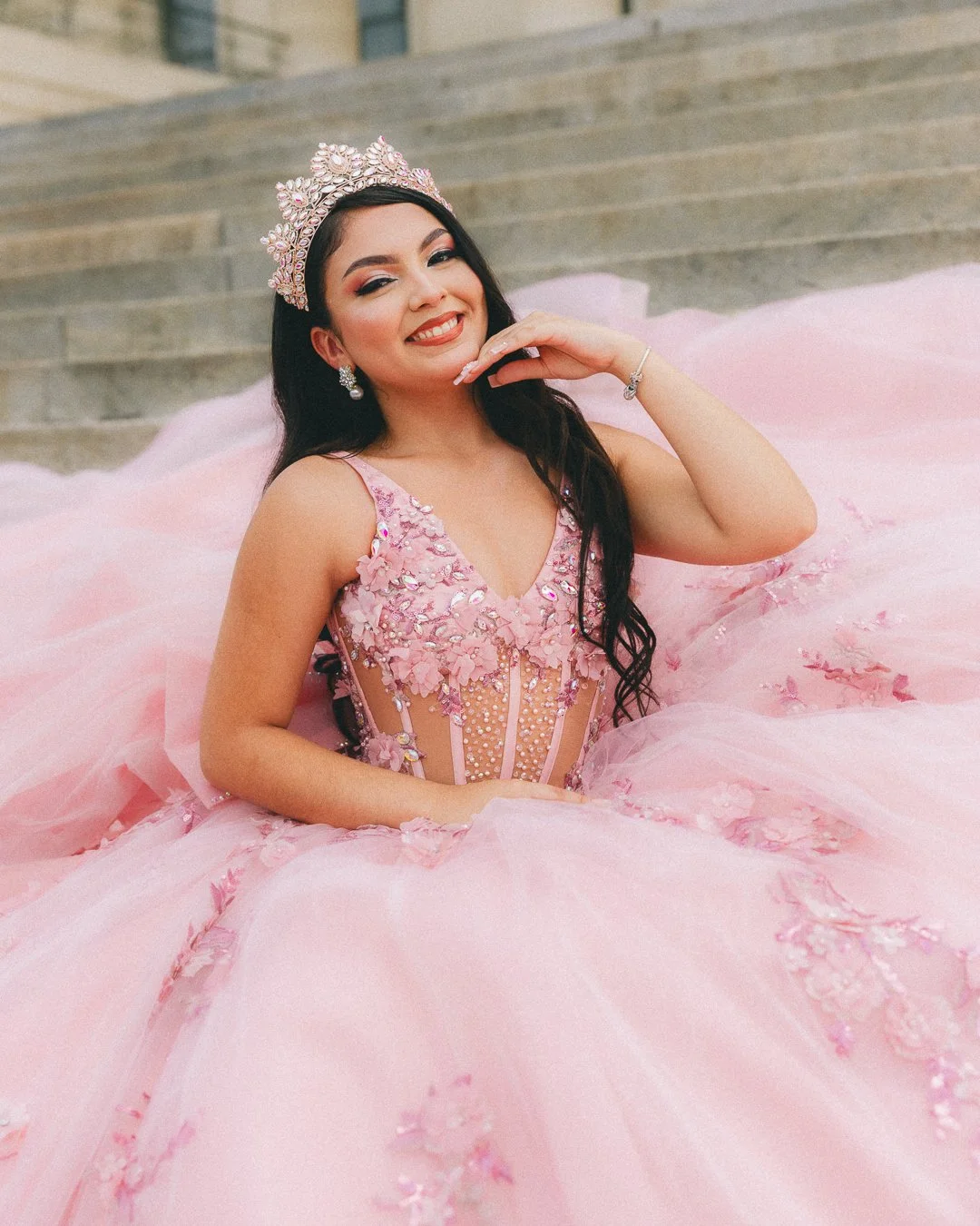 Young woman in a pink ball gown with floral embellishments and a tiara, sitting on steps outside, smiling and posing with her hand near her face.
