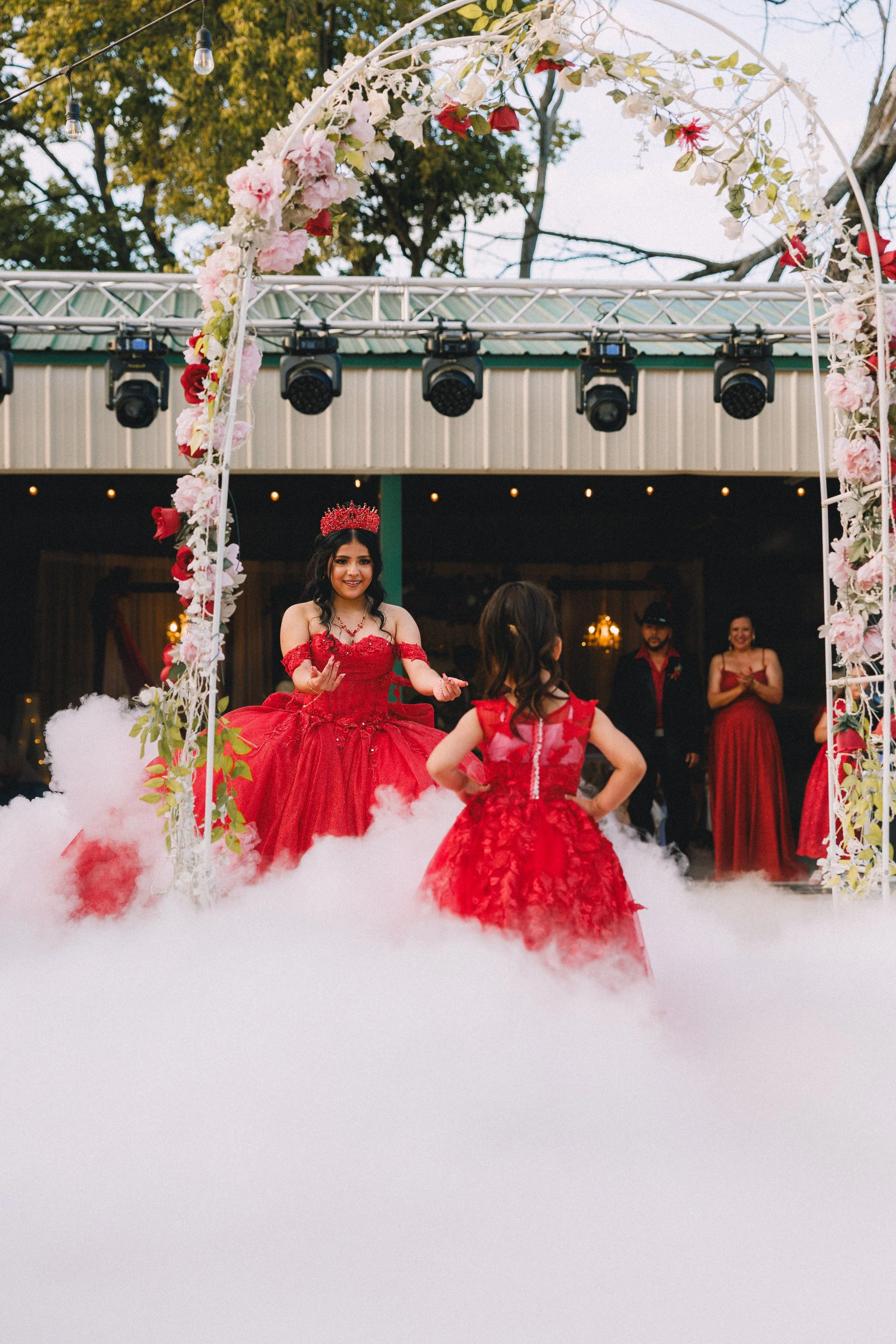 A young woman dressed as a queen wearing a red dress and a crown, dancing with a young girl in a red dress at a celebration or party, with a decorated arch and onlookers in the background.