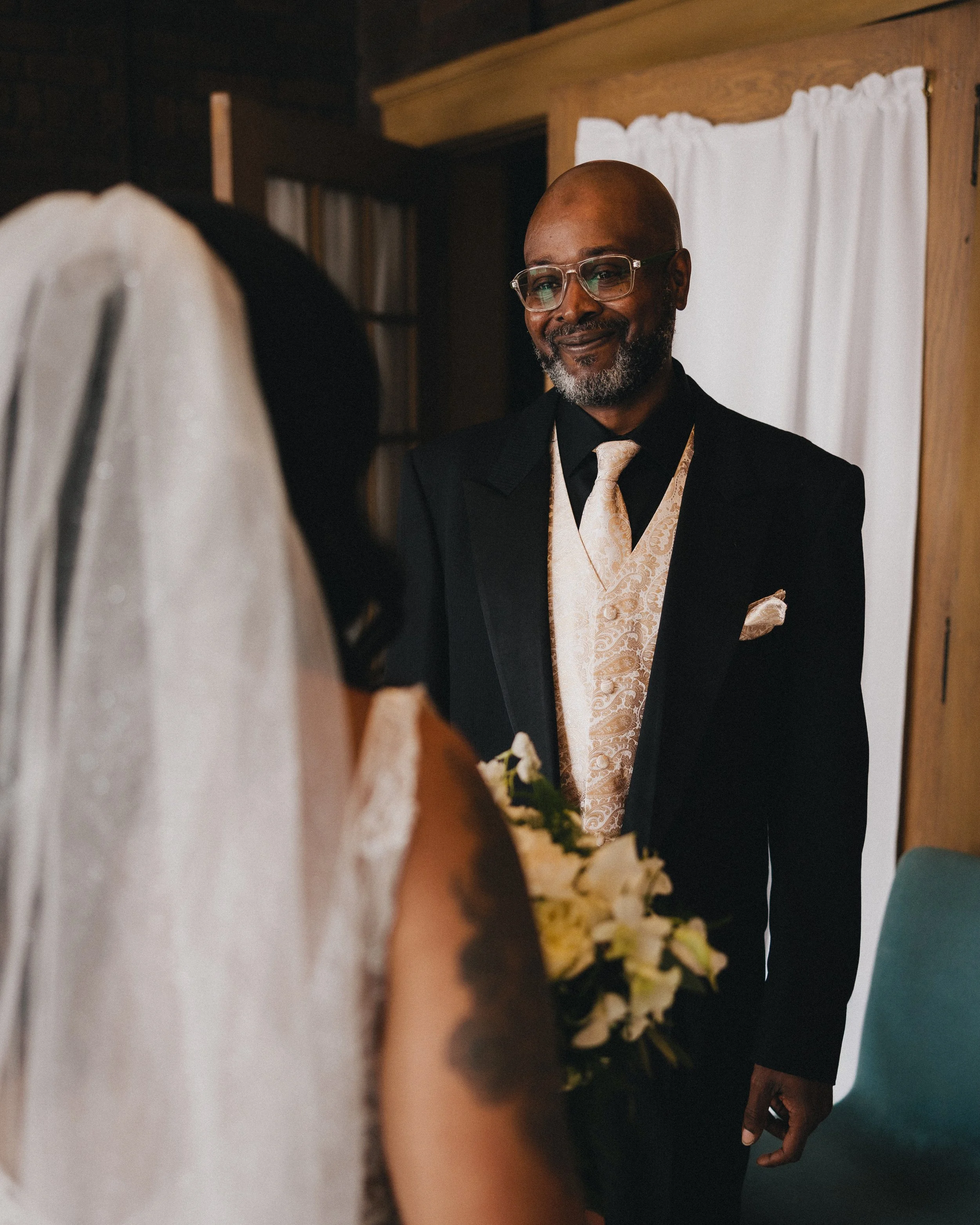 A man dressed in a black tuxedo with a gold patterned vest, wearing glasses, smiling and holding a bouquet of white flowers, standing in front of a white curtain. Shot at Topeka Kansas venue The Beacon.