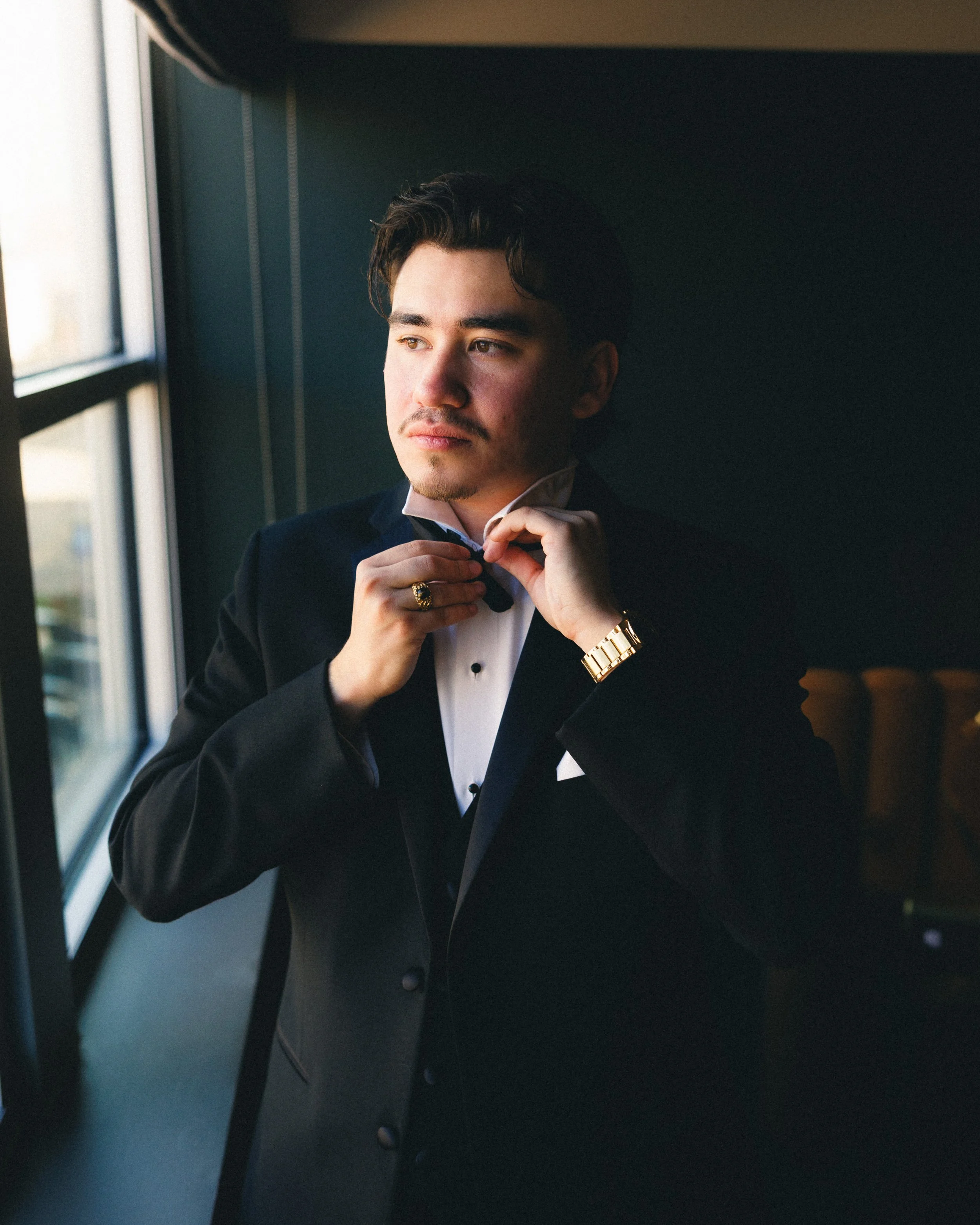 Young man in formal black tuxedo adjusting his bowtie by a window. Shot at Cyrus Hotel in Topeka, Kansas. 
