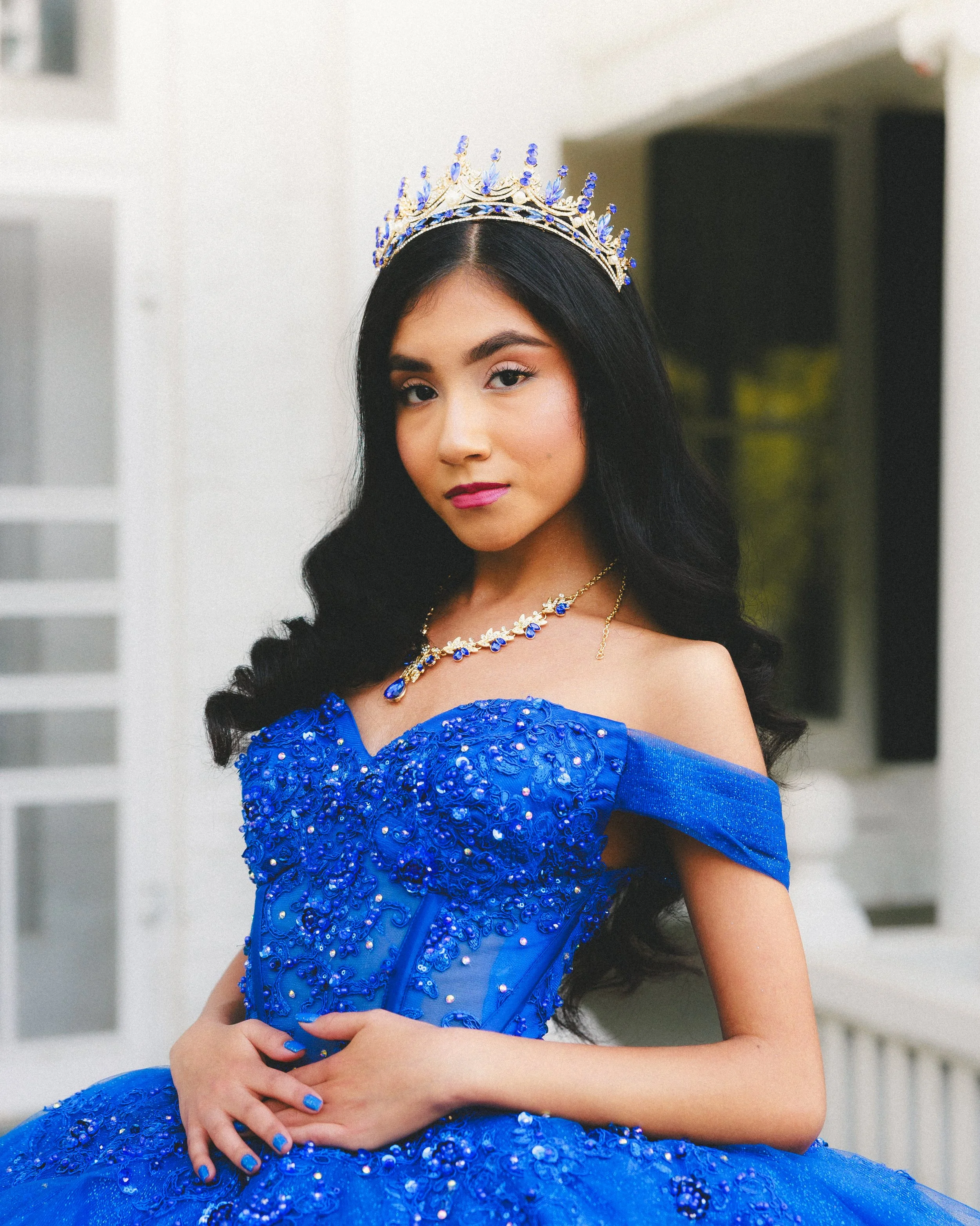 Young woman wearing a blue ballgown with beadwork, a tiara, and matching jewelry, standing with hands crossed in front of a white background.