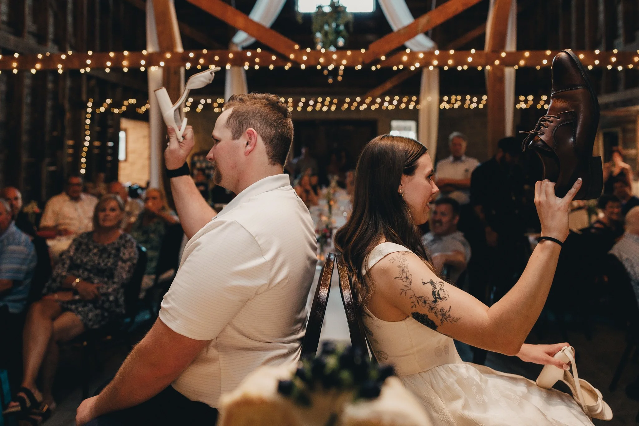 A man and woman sitting back-to-back at a rustic wedding reception, participating in the shoe game, with guests watching and string lights hanging above.