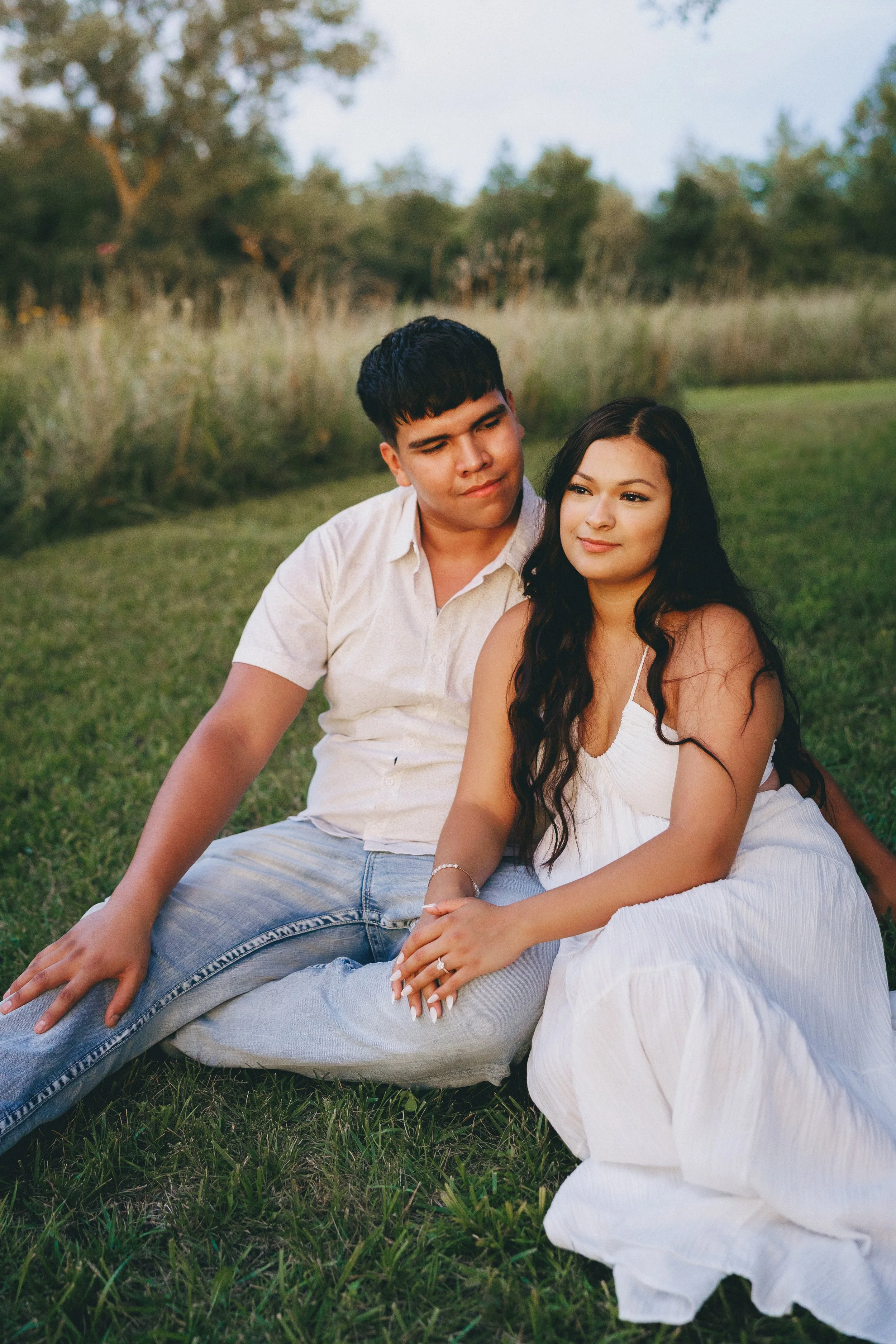 A young couple sitting on grass in a park with trees in the background during the evening.  Shot in Topeka, Kansas. 