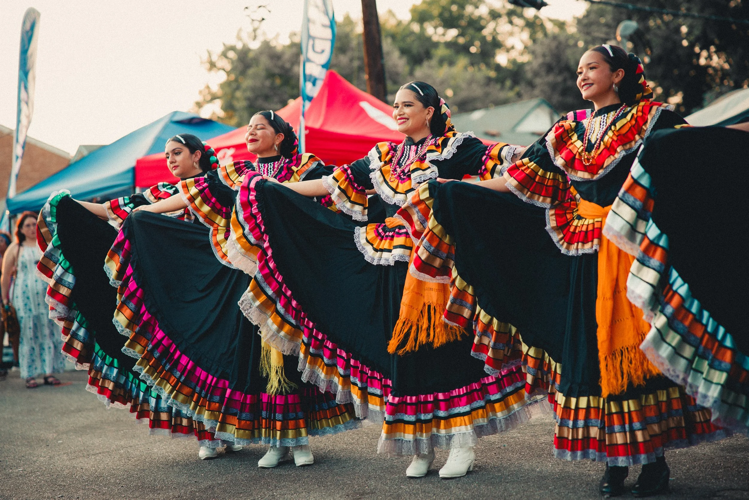 Five women dressed in colorful traditional Mexican folkloric dresses perform a dance outdoors at a cultural festival. Shot at Fiesta Topeka.