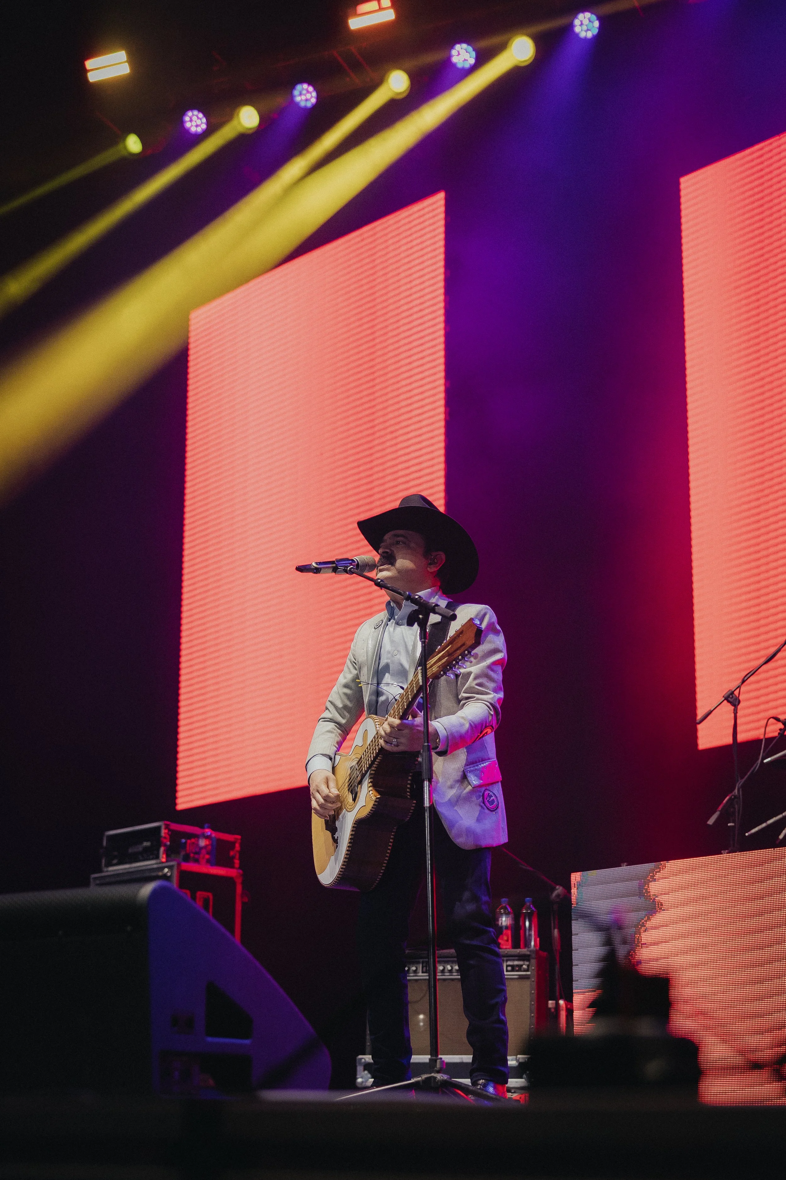 A musician with a cowboy hat and gray blazer is playing an acoustic guitar and singing into a microphone on stage with red and purple LED background lights. Los Tucanes de Tijuana Stormont Vail Event Center Topeka, Kansas. 
