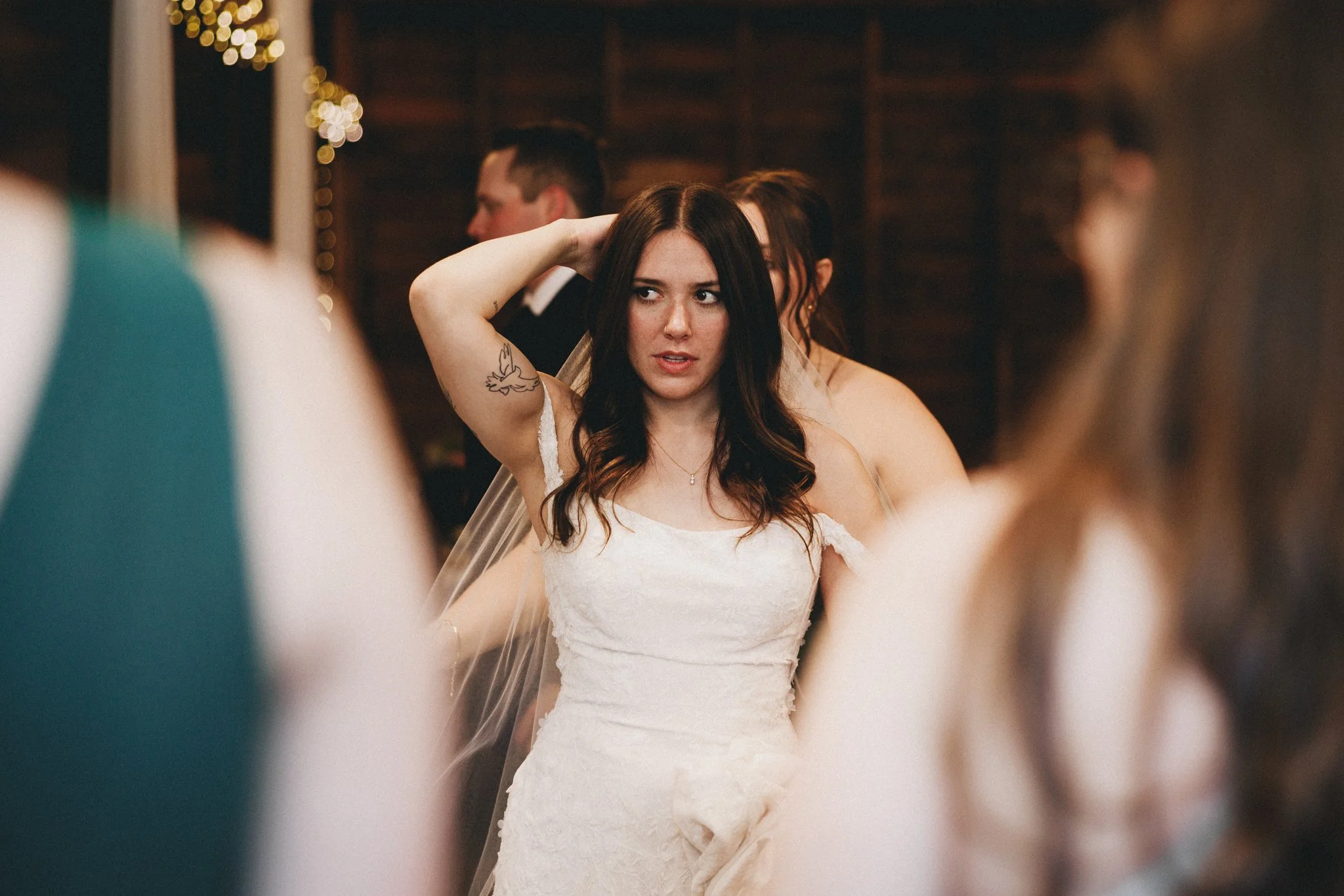 A woman in a wedding dress with dark hair stands among other wedding guests during a reception, adjusting her hair while looking to the side.