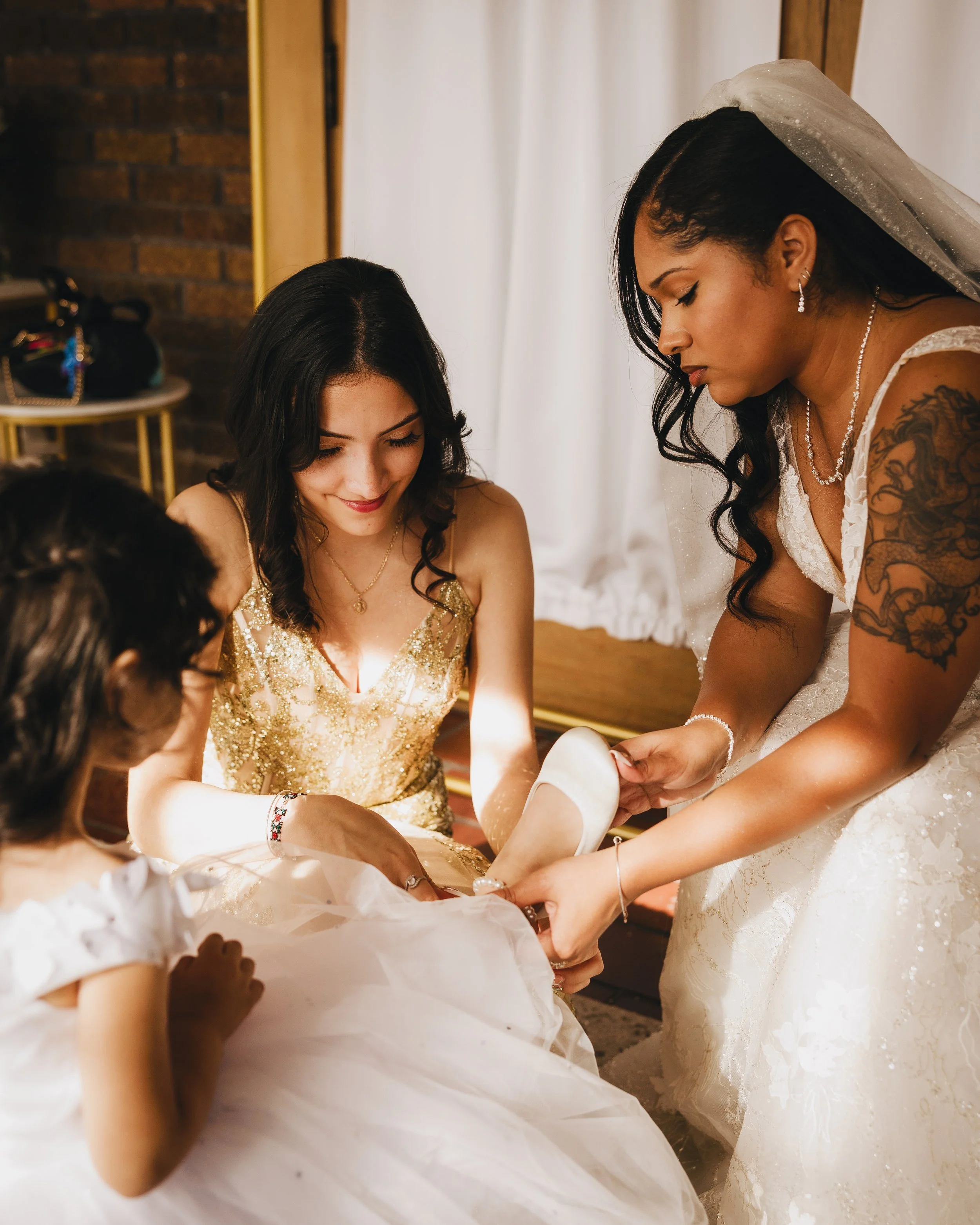 A bride putting on her white wedding shoe with the help of a woman in a gold dress and two young girls in white dresses. Shot at Topeka Kansas venue The Beacon.