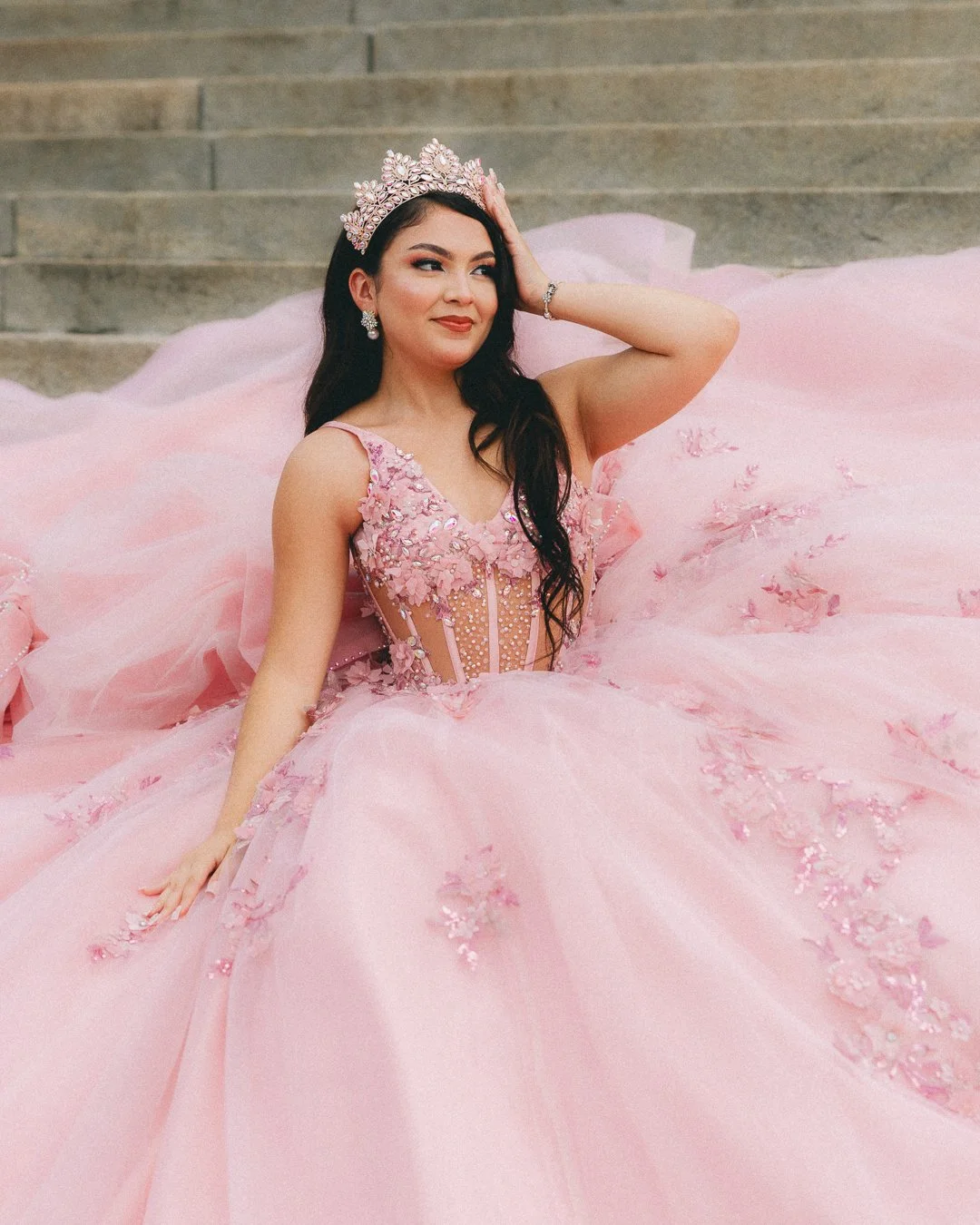 A woman wearing a pink quinceañera dress with floral embroidery and sequins, sitting on stairs, wearing a tiara, earrings, and a bracelet.