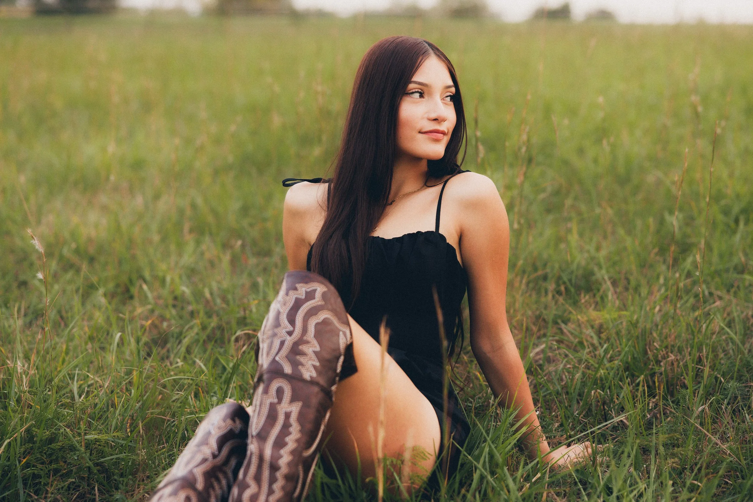 A young woman with long dark hair sitting in a grassy field, wearing a black dress and patterned boots.