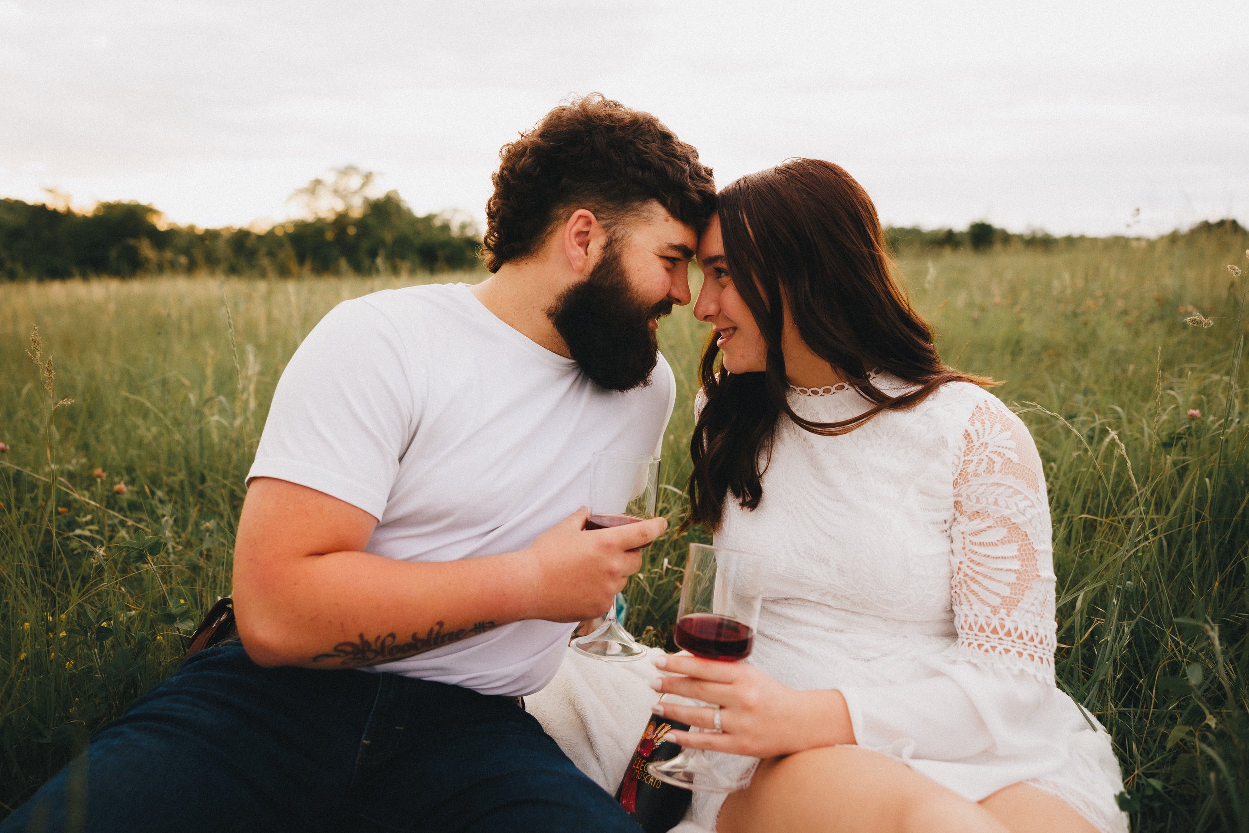 A couple sitting in a field of grass, touching foreheads and smiling while holding glasses of red wine.