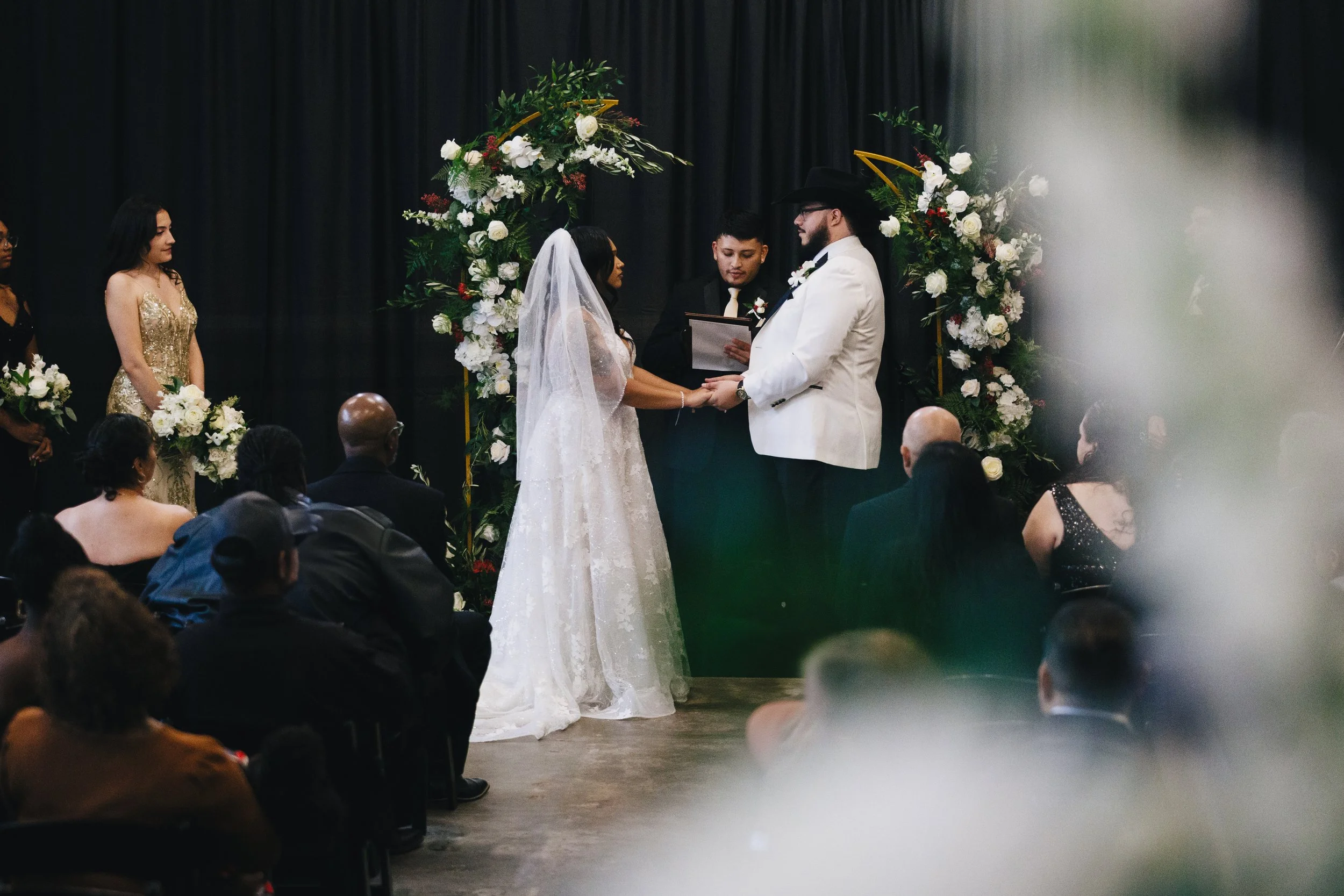 A wedding ceremony with a bride and groom holding hands, standing in front of a floral arch, surrounded by guests seated and standing, with a black curtain backdrop. Shot at Topeka Kansas venue The Beacon.