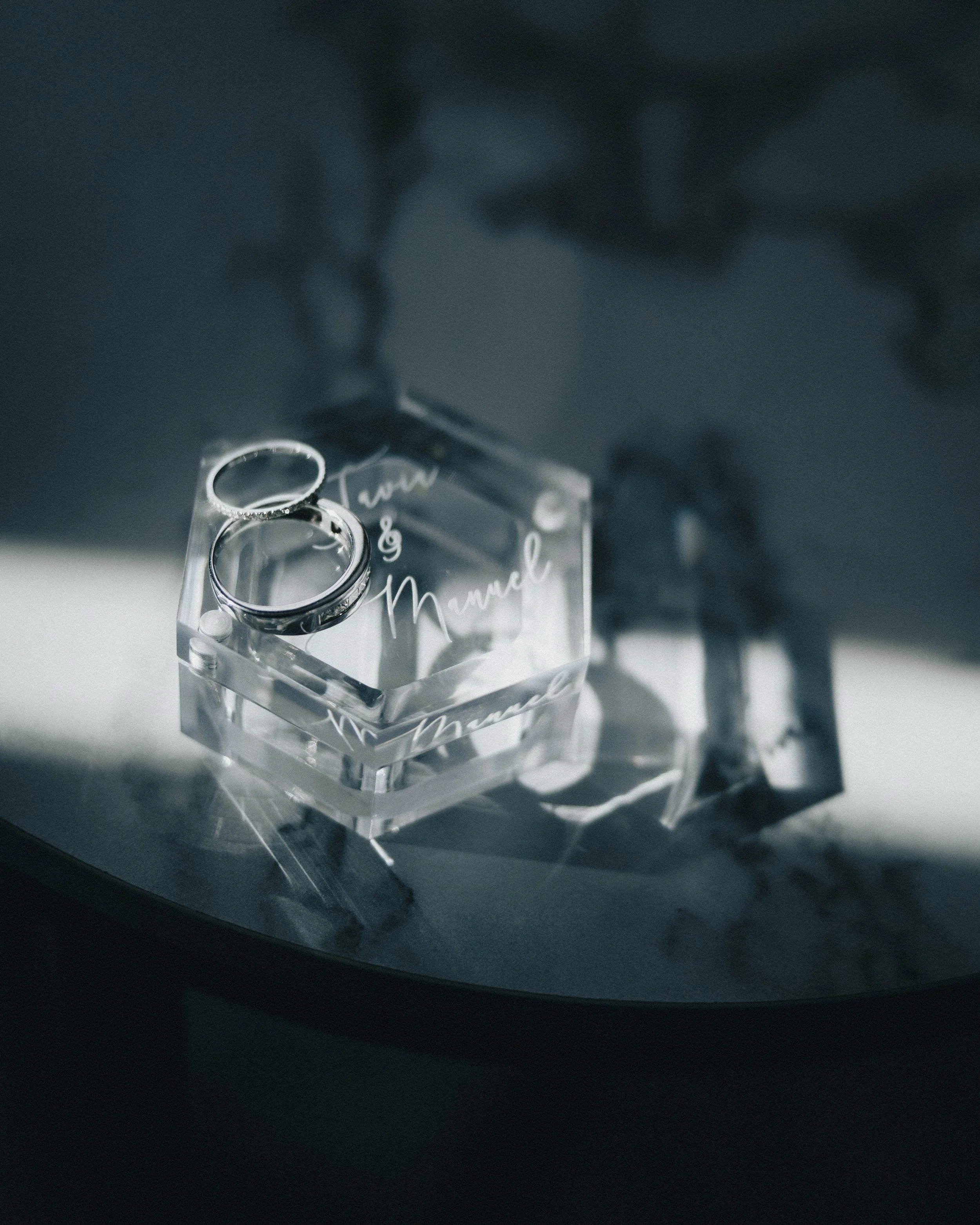 A pair of silver rings on a clear acrylic stand with the words 'Love & Mua' written on it, placed on a reflective surface in a dimly lit setting. Shot at Topeka Kansas venue The Beacon.
