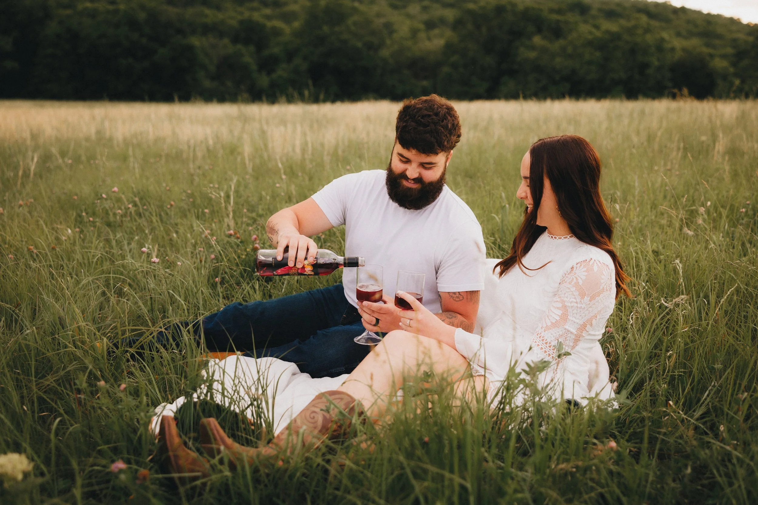 A man and woman sitting in a grassy field, with the man pouring red wine into the woman's glass, both smiling and enjoying a picnic outdoors.