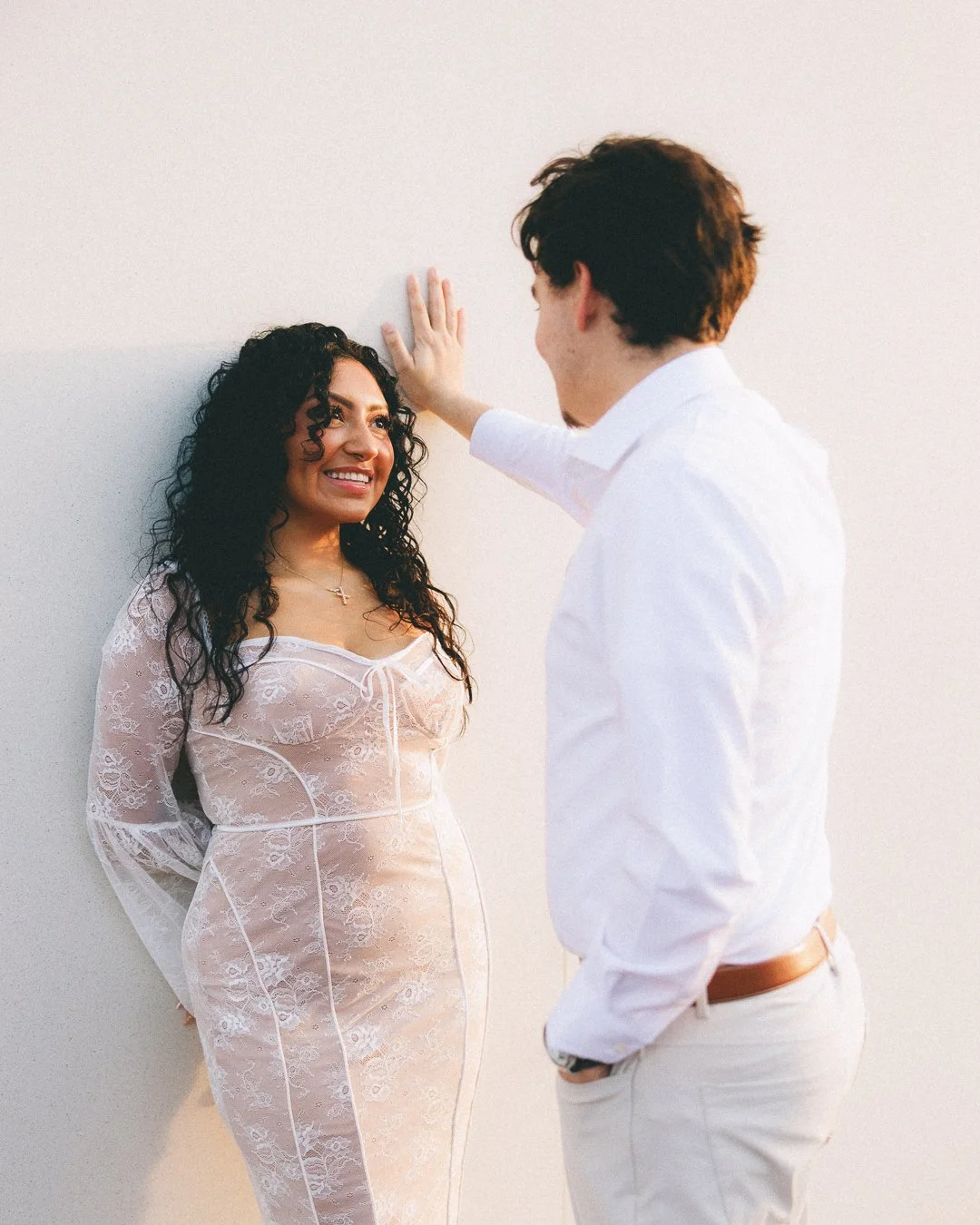 A woman with curly black hair smiling at a man with brown hair. She is wearing a sheer lace dress and a cross necklace. The man, dressed in a white shirt and beige pants, is leaning against a plain white wall, with his hand touching the wall near her