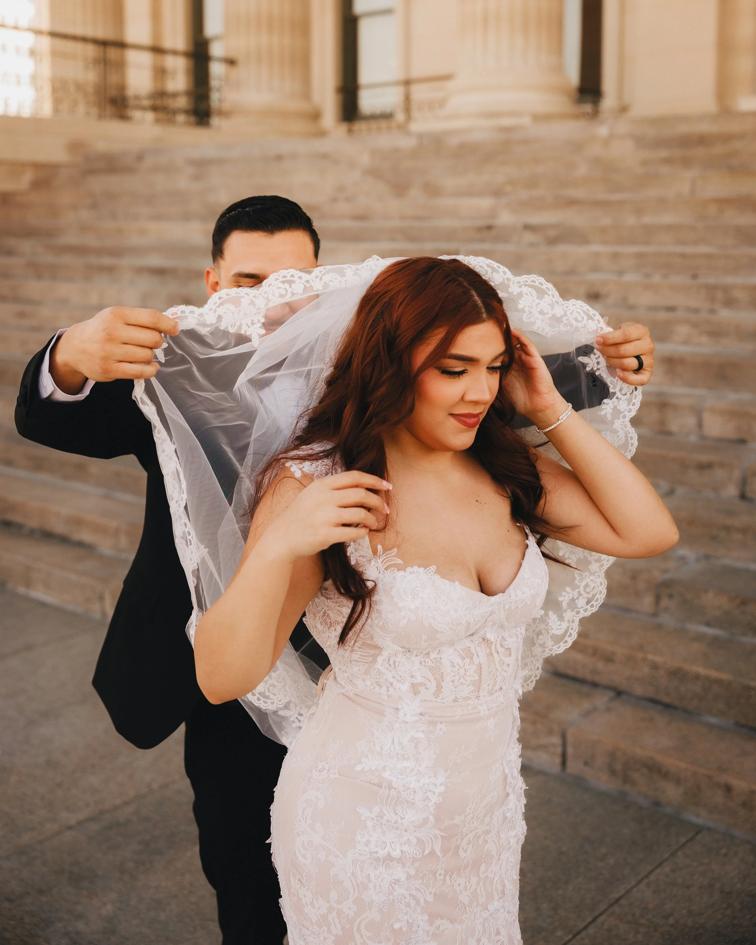 A bride with long red hair in a white lace wedding dress, adjusting her veil, with a groom in a black suit behind her, on stone steps outside a building.