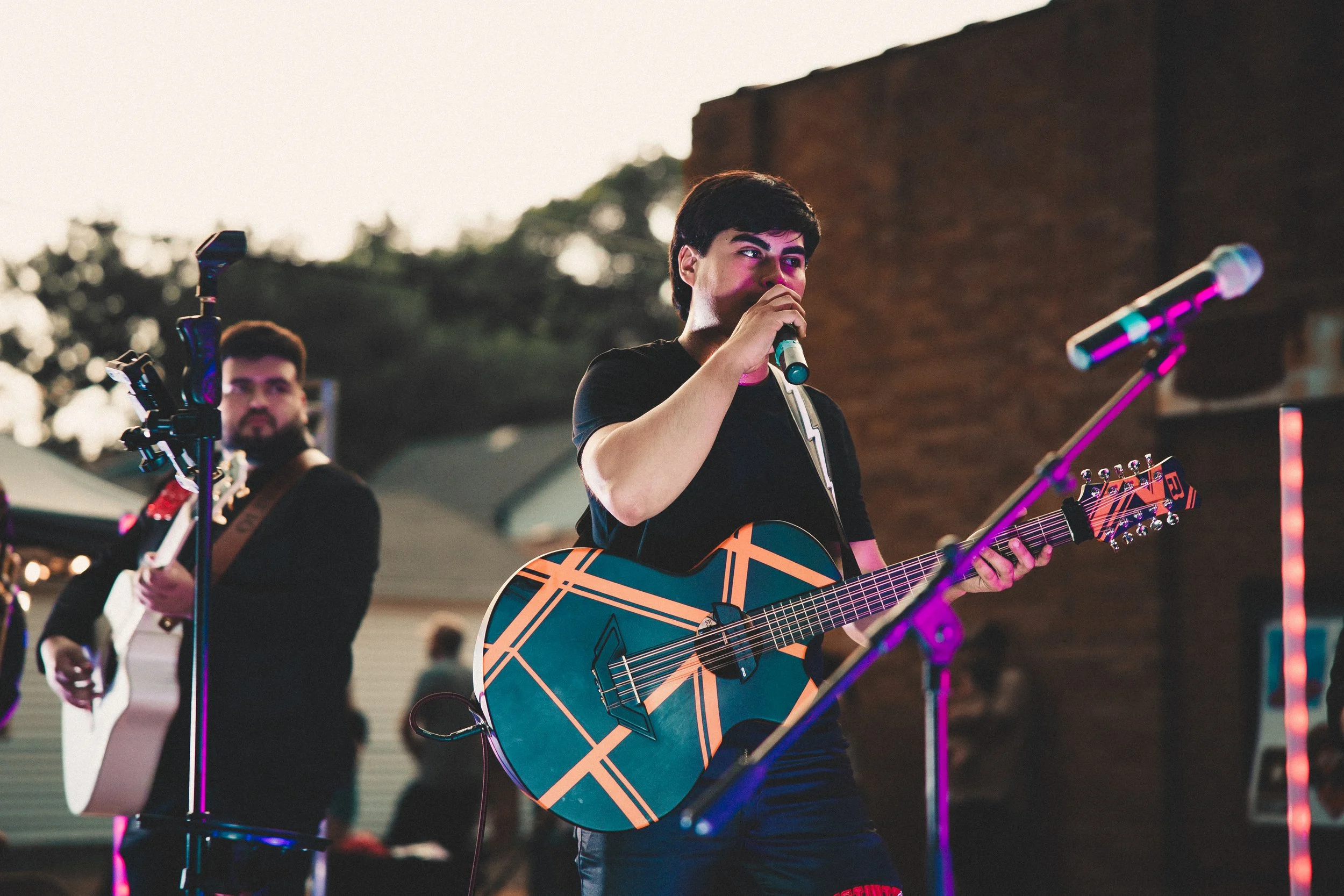 Young man singing into a microphone while playing an acoustic guitar on an outdoor stage, with another guitarist in the background. Shot at Fiesta Topeka.