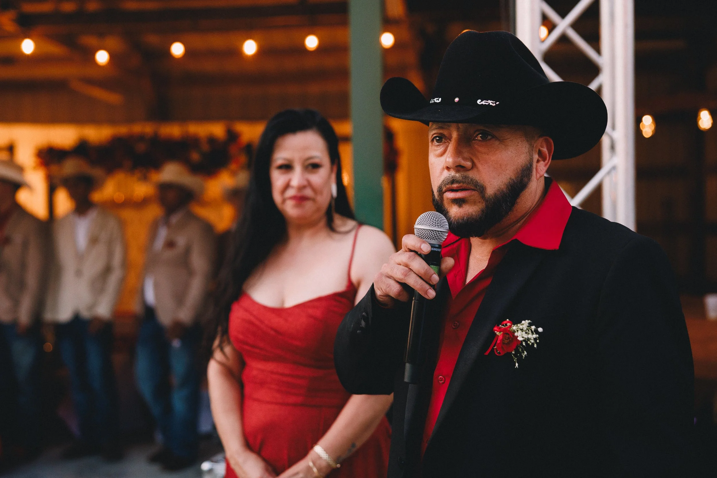 A man wearing a black cowboy hat, red shirt, and black blazer speaking into a microphone at a formal event. A woman in a red dress stands beside him, looking on. In the background, there are blurred people and decorative lights.