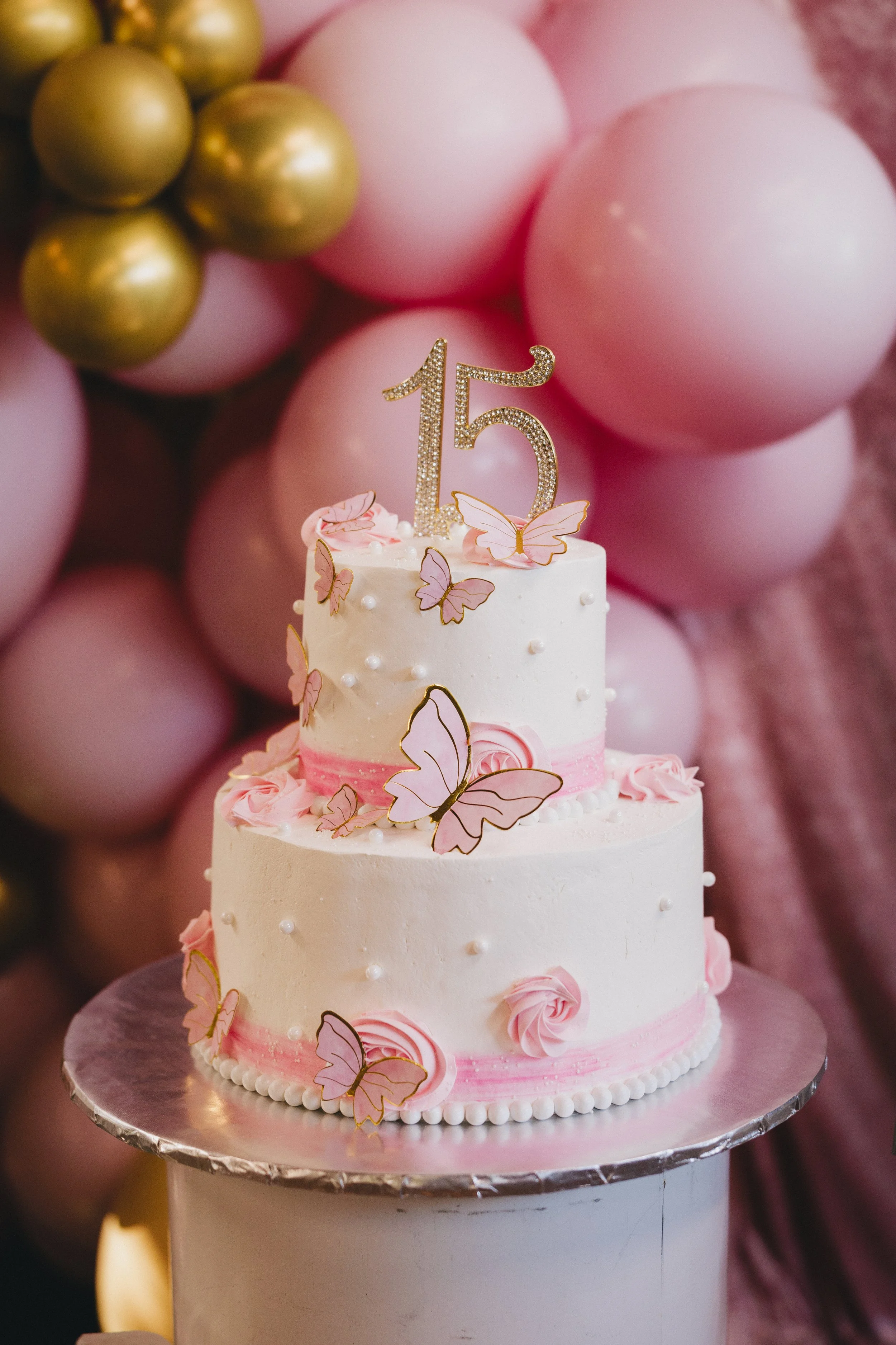 A two-tiered birthday cake decorated with pink and white frosting, pink rosettes, pearl-like beads, and butterfly toppers, with a glittery number 15 candle on top, against a backdrop of pink and gold balloons.