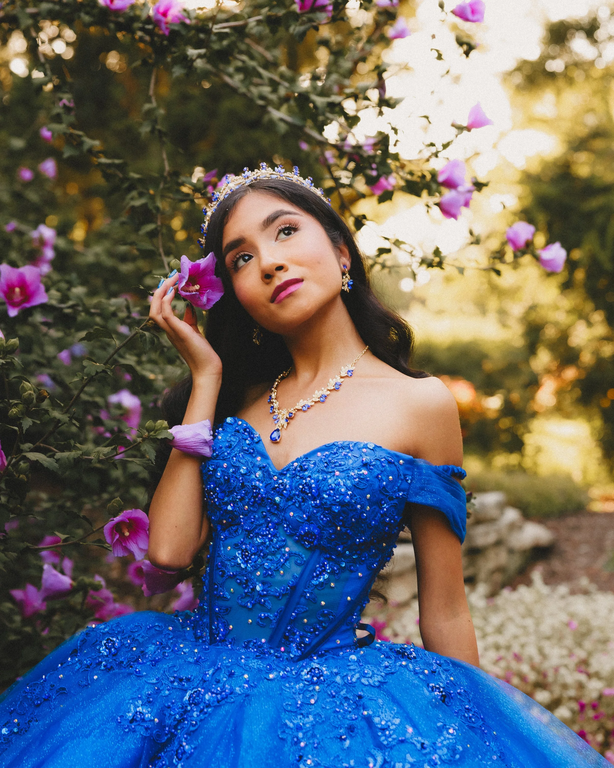 Young woman in an elegant, royal blue, off-shoulder gown with intricate beadwork, sitting among pink flowers and greenery, wearing a matching jewelry set and a tiara, in a garden setting with sunlight filtering through trees.