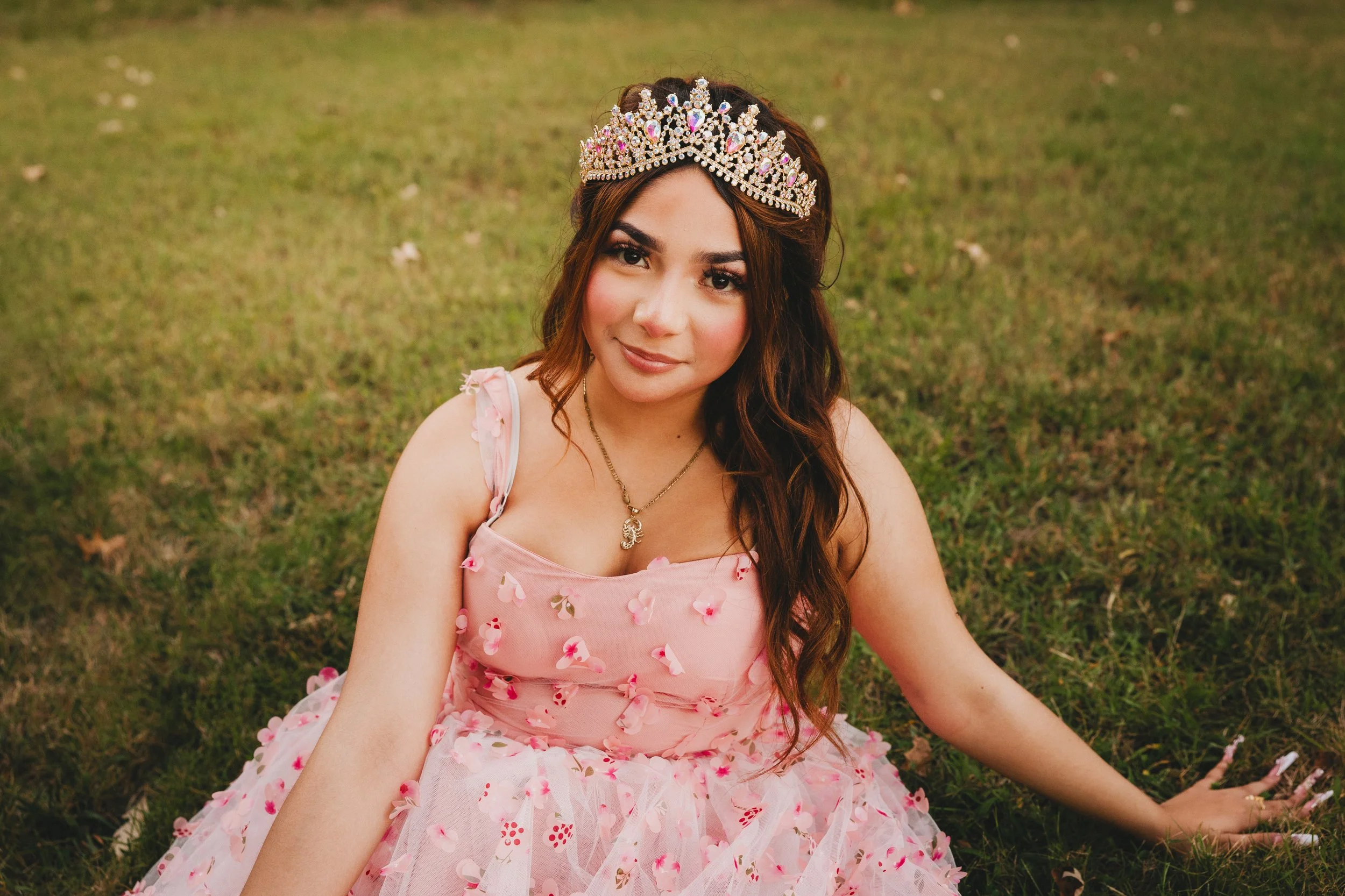 Young woman sitting on the grass, wearing a pink dress adorned with flower petals and a tiara, smiling gently.