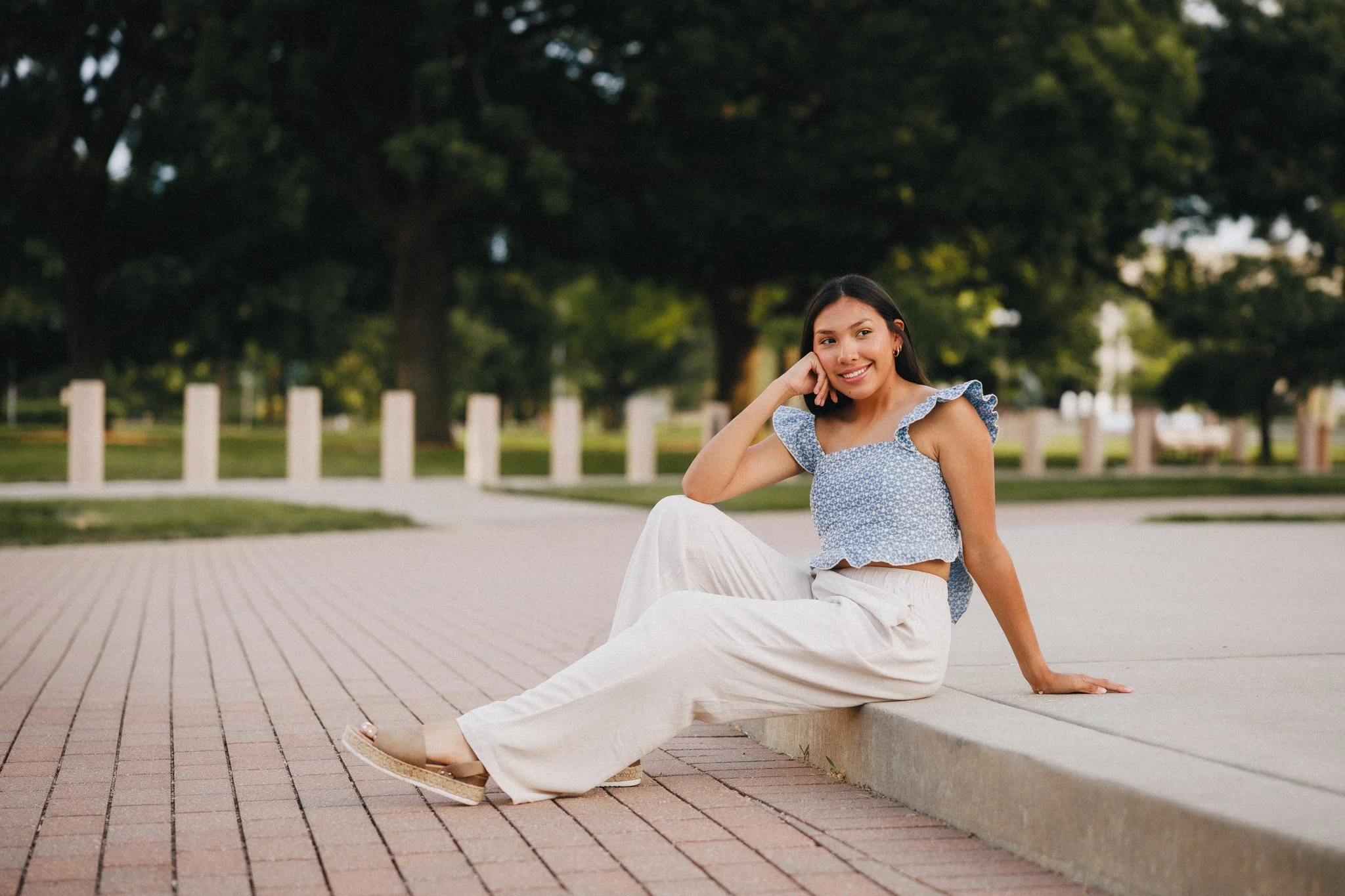 A young woman sitting on a concrete ledge on a paved walkway in a park, smiling at the camera with trees in the background.