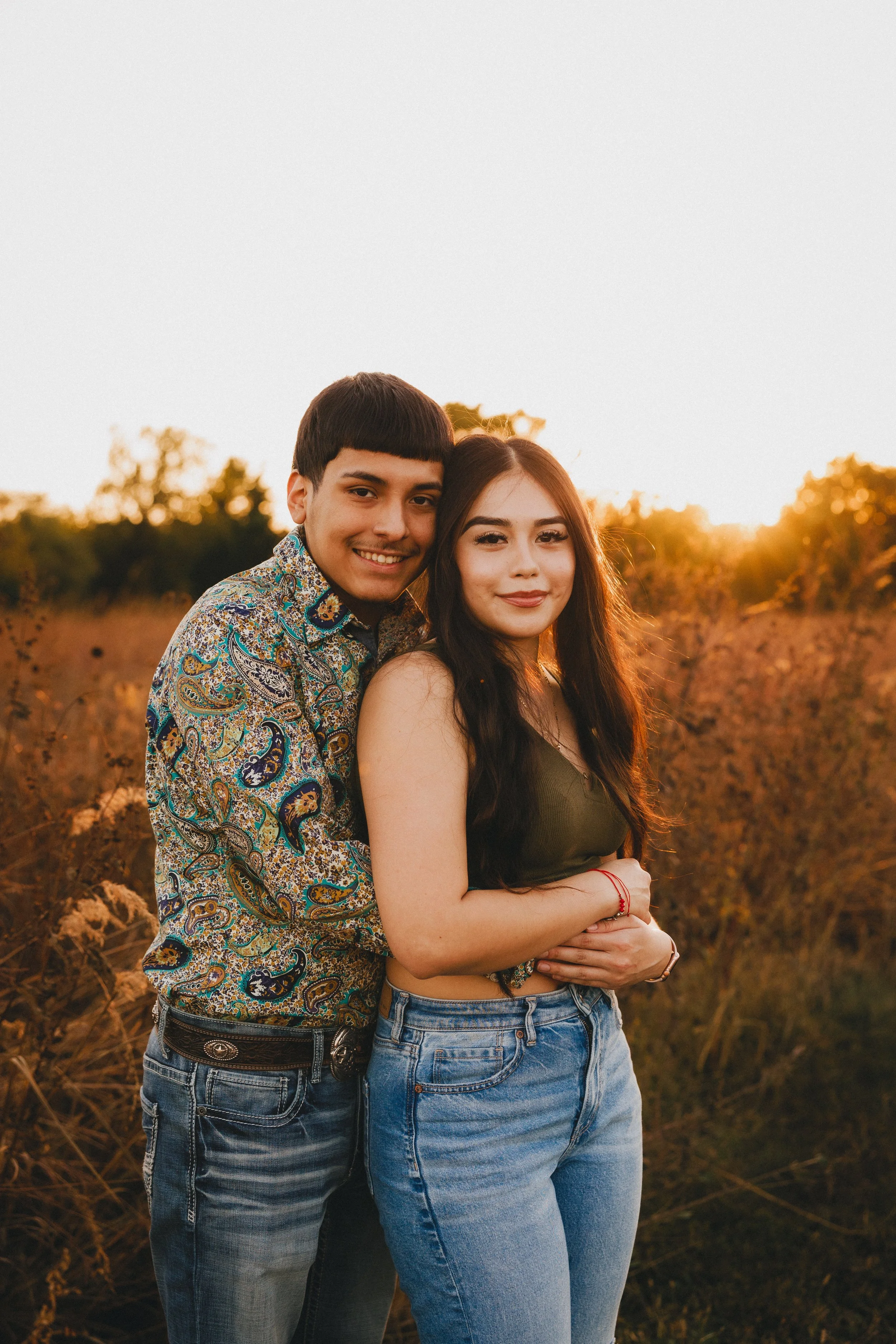A young couple standing closely together outdoors at sunset, smiling and embracing in a field of tall dry grass and bushes. Shot in Topeka, Kansas. 