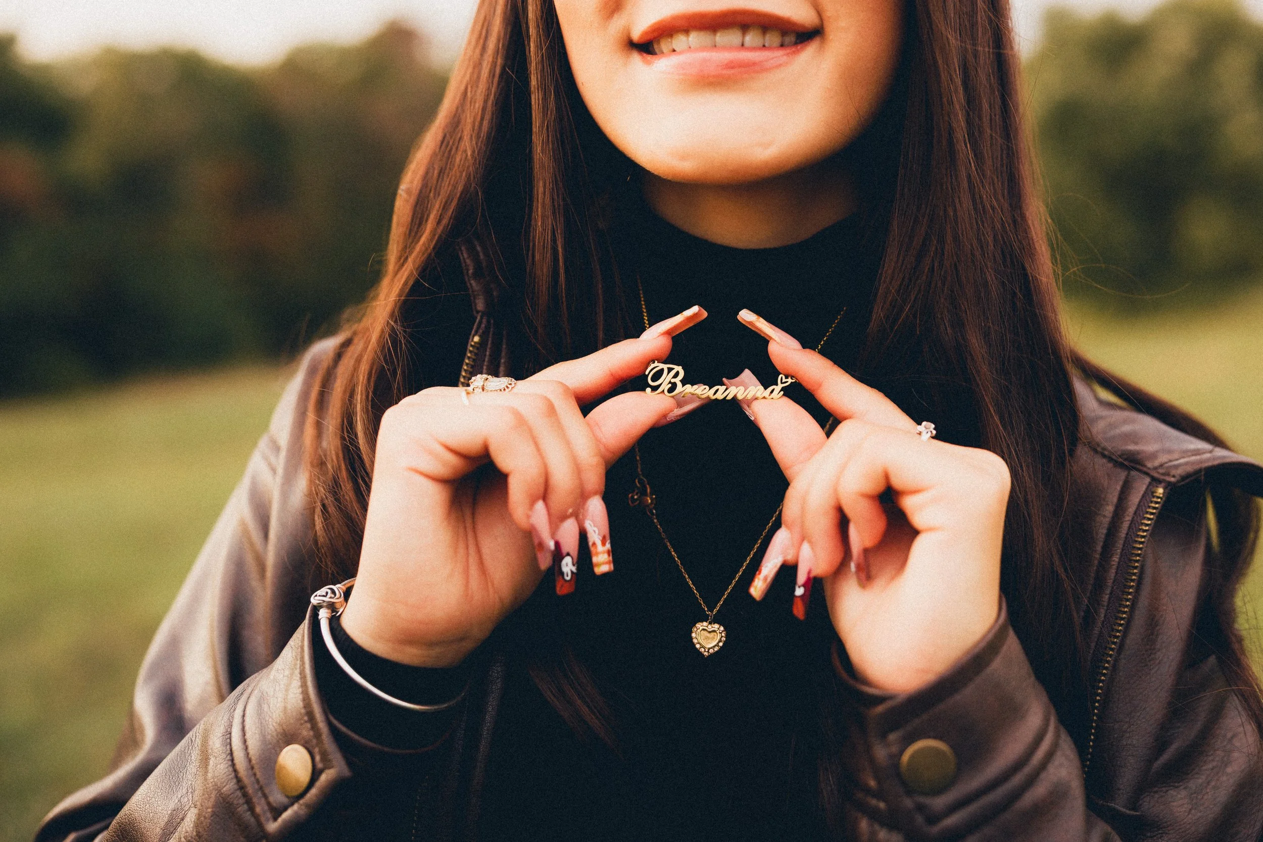 Close-up of a woman holding a necklace with the word 'Breanna' in gold, smiling with visible teeth, outdoors with blurred green background.