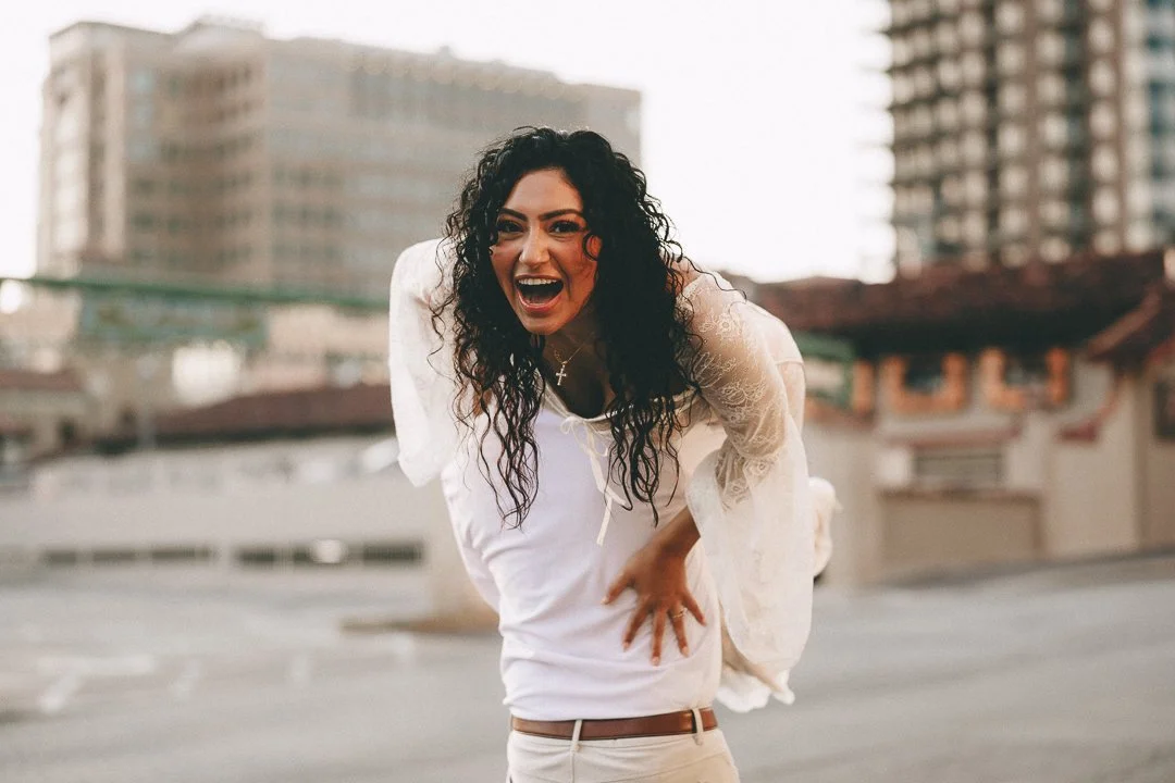 Woman with curly hair wearing a white top, laughing outdoors in an urban area with high-rise buildings in the background. Shot at Kansas City, Country Club Plaza.