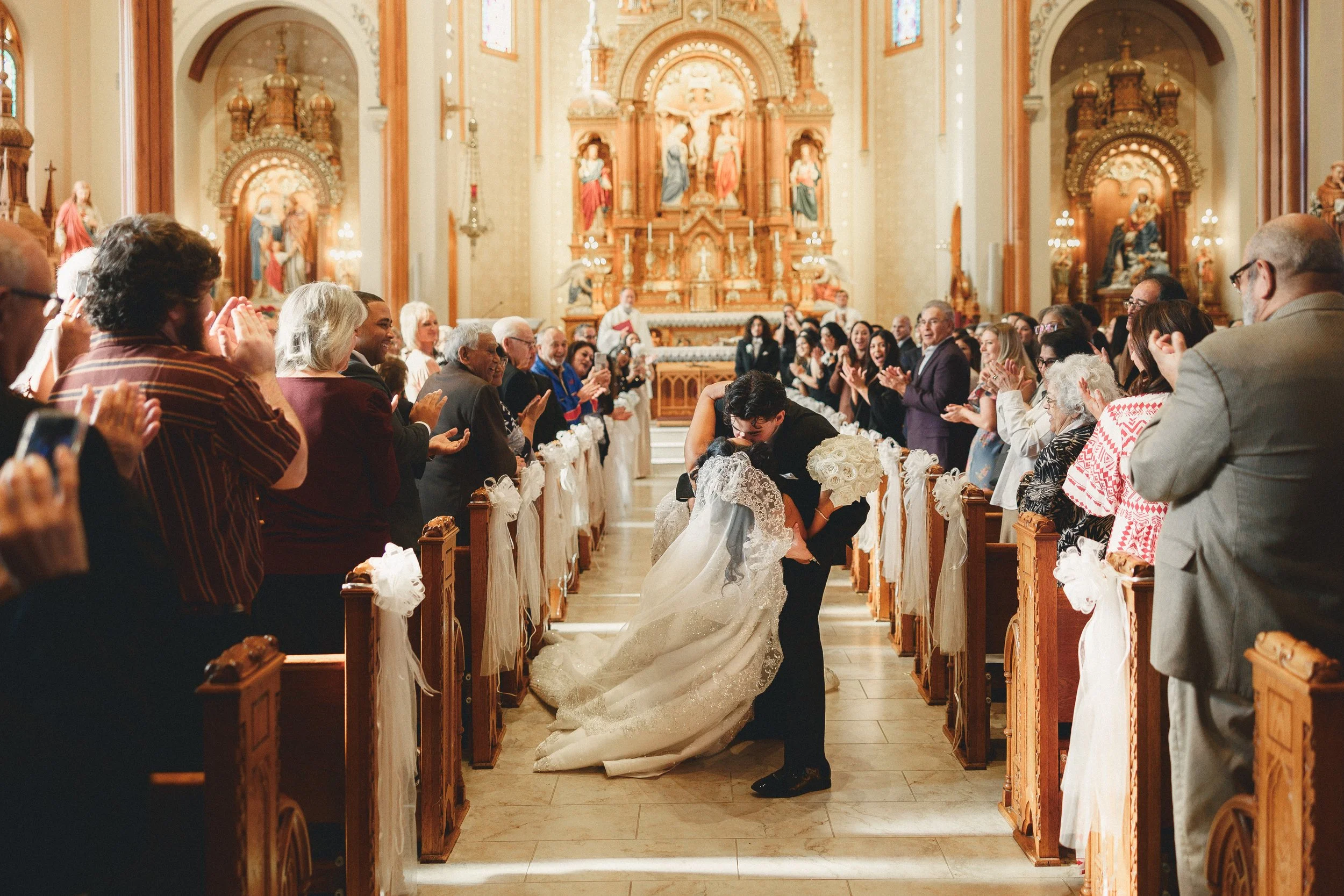 A newlywed couple is kissing in a church aisle, surrounded by cheering guests during their wedding ceremony.