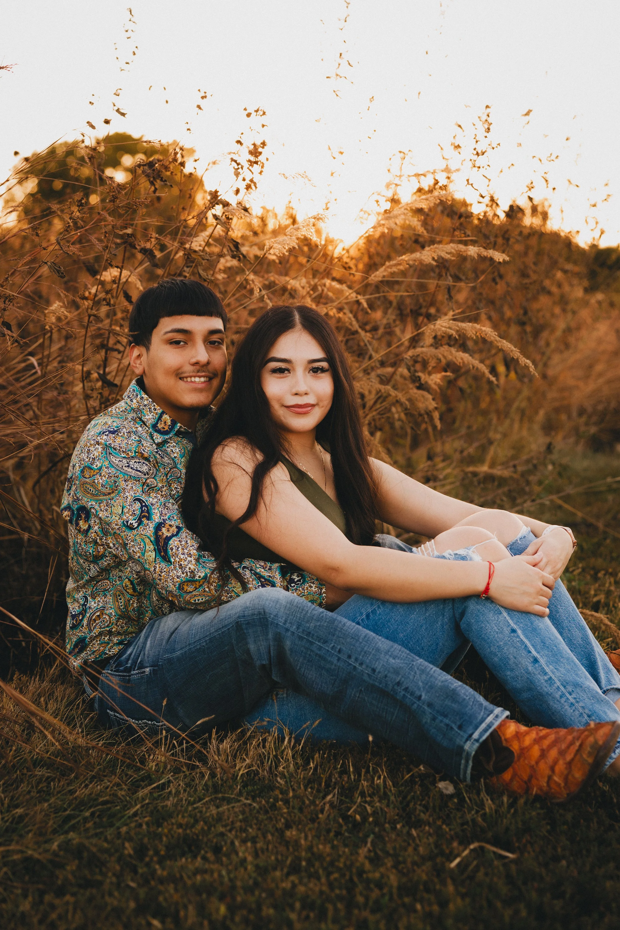 A young couple sitting together on the grass during sunset, with tall, dry grass behind them and a warm, orange sky in the background. Shot in Topeka, Kansas. 