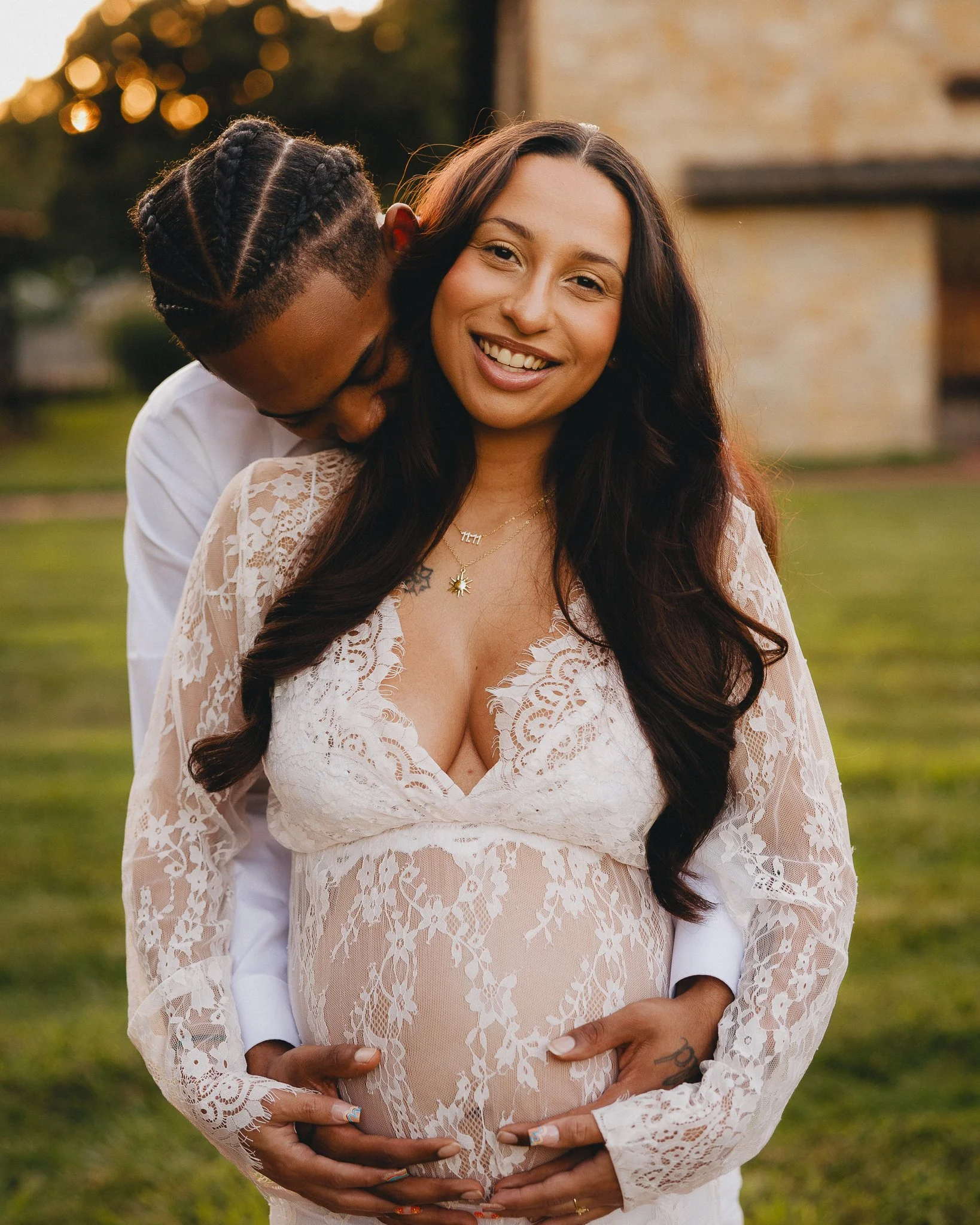 A pregnant woman with long dark hair in a lace dress faces the camera, smiling, with a man kissing her on the side of her head from behind. The man has braided hair and is wearing a white shirt. They are outdoors on a grassy area with trees and a sto