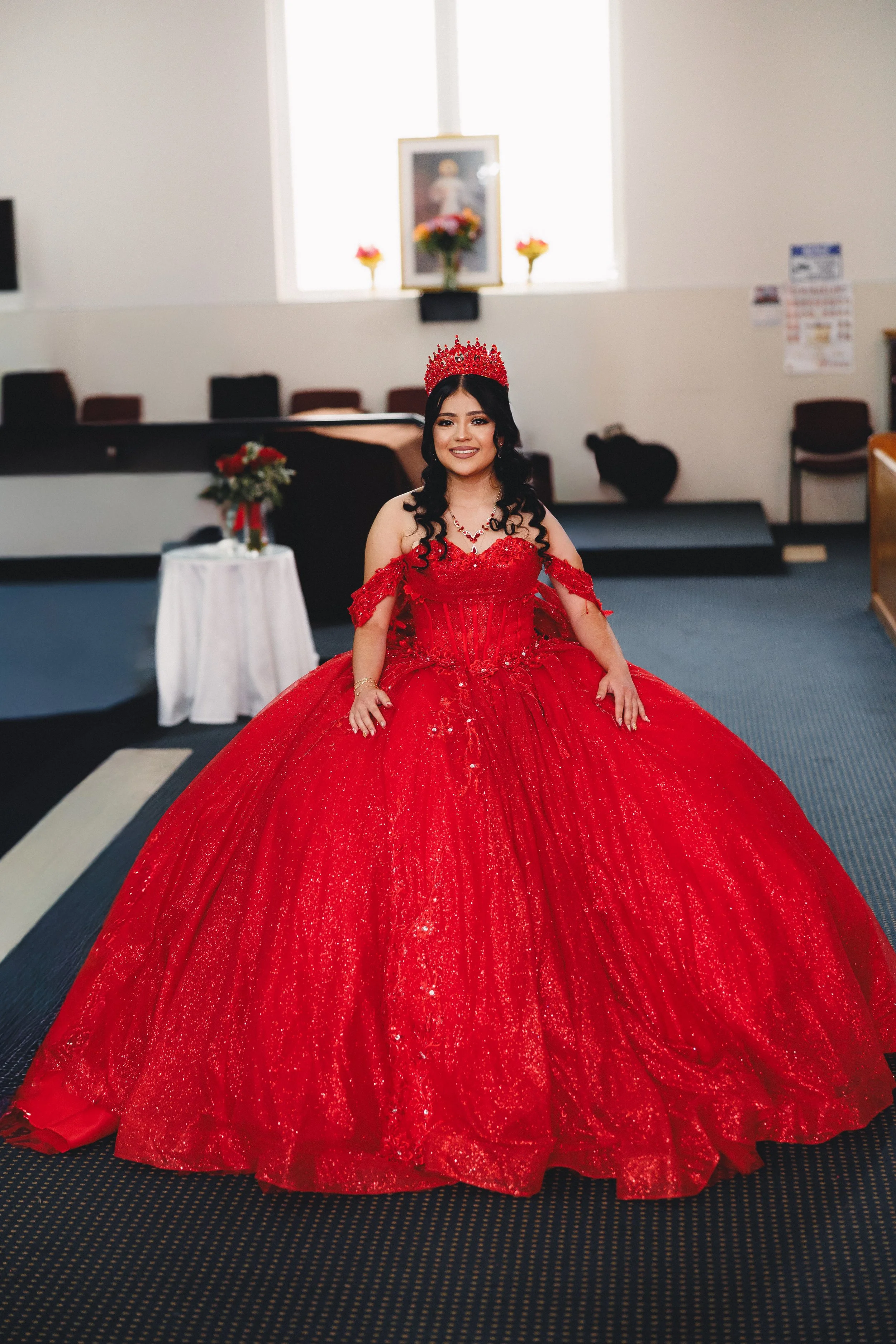 A woman wearing a vibrant red, sparkling ball gown with off-shoulder lace sleeves, a matching crown, and jewelry, standing inside a room with a religious picture and flower arrangements in the background.