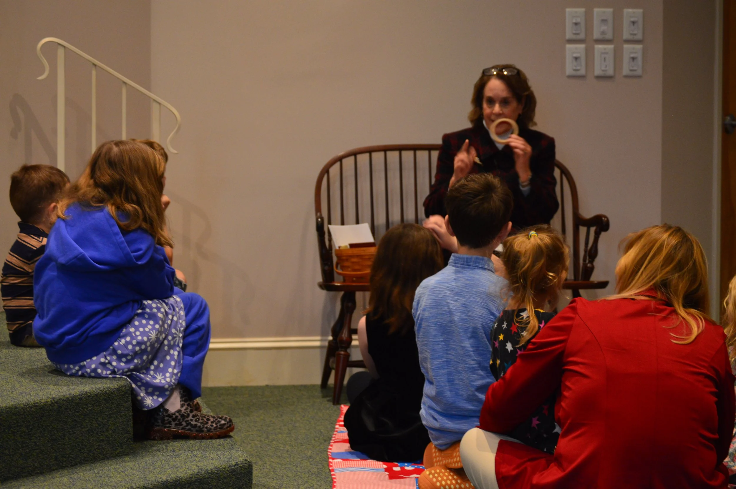 A group of children sitting cross-legged on a colorful rug, listening attentively to a woman who is speaking to them and holding a circular object. The children are in our sanctuary participating in children's time during a service.