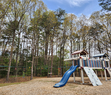 Children's playground with a blue slide, wooden climbing structure, and trees in the background on a gravel surface.