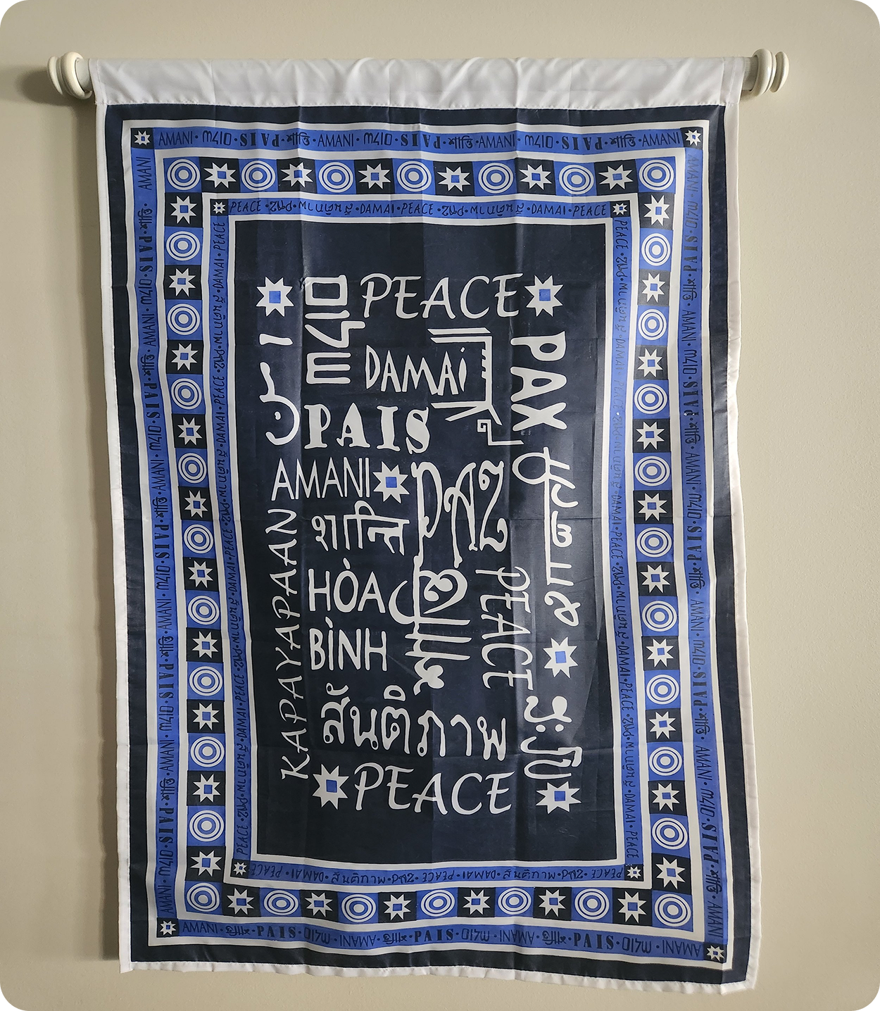A fabric wall hanging with the phrase "Peace in my homeland, Peace in my country" written in various languages, framed by a pattern of stars and circles in blue, white, and black colors.