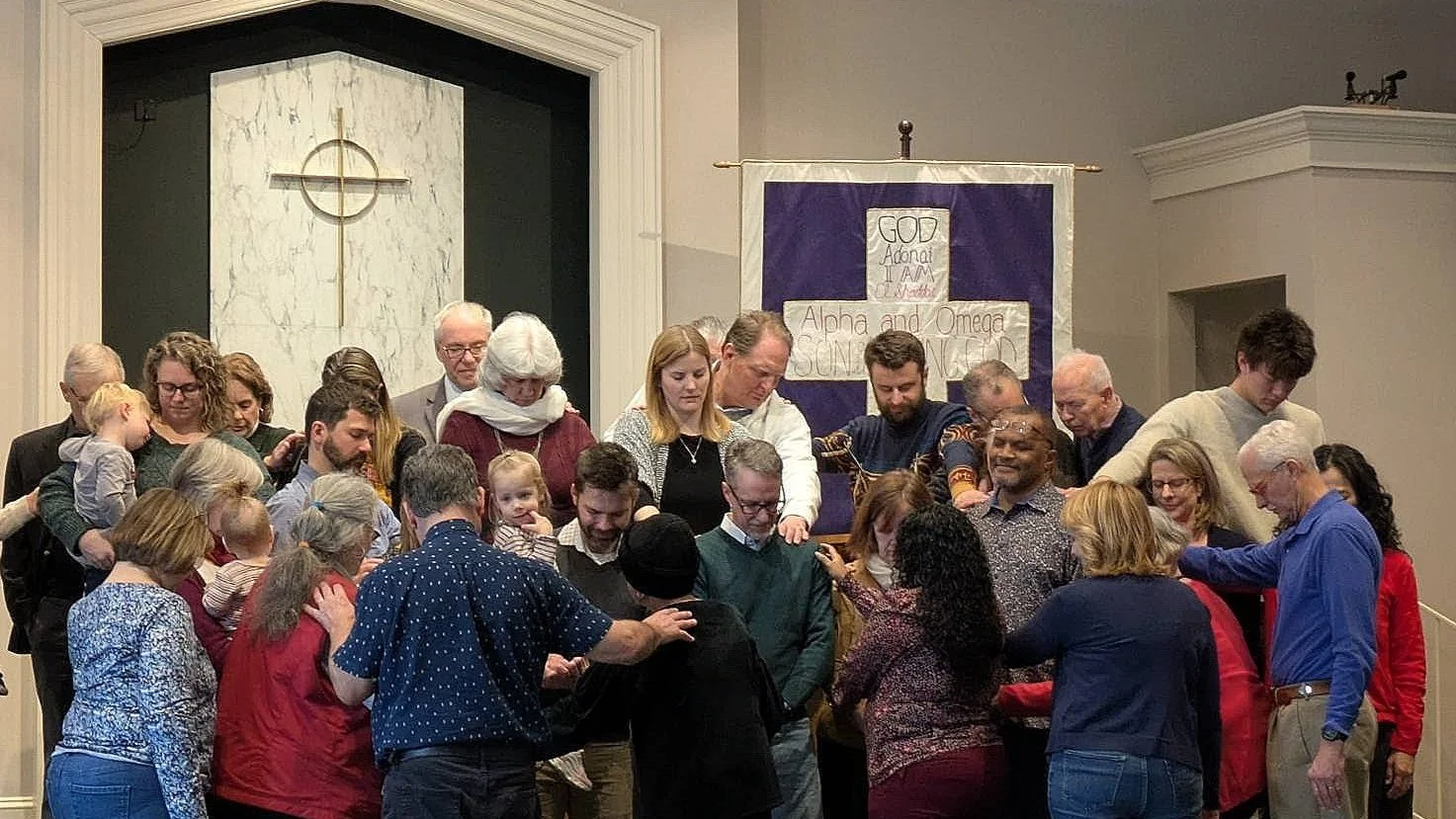 A diverse group of people gathered in prayer inside a church near a cross on the altar with a purple and white banner in the background.