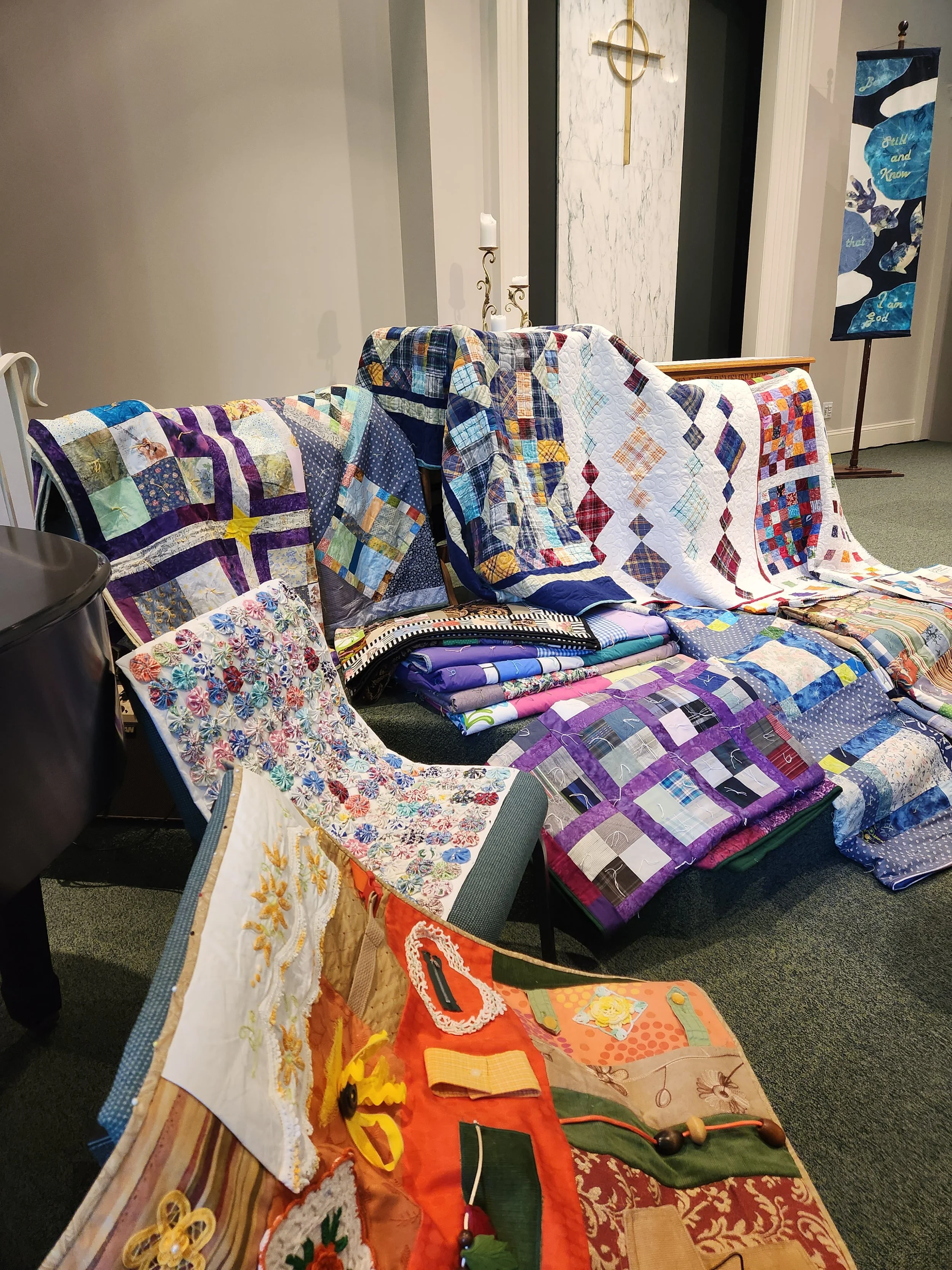 Various colorful quilts and fabric pieces arranged on tables and the floor, with some decorative elements and fabric craft supplies visible.